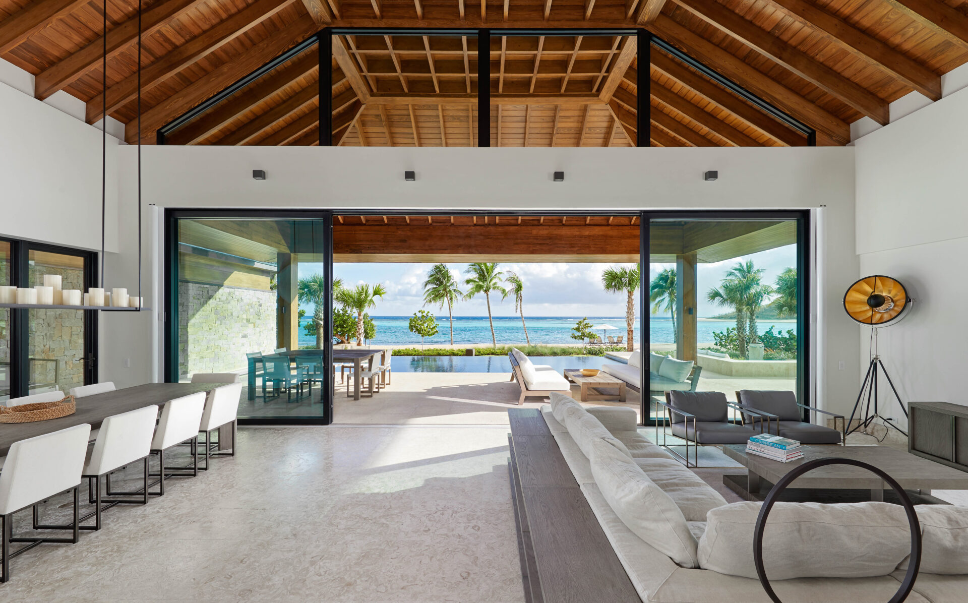 A spacious, modern living area with high wooden ceilings and open sliding glass doors revealing a scenic ocean view. The room features white sofas, a long dining table with white chairs, an outdoor seating area, palm trees, and a clear blue sky – the perfect setting for a seaside vacation.