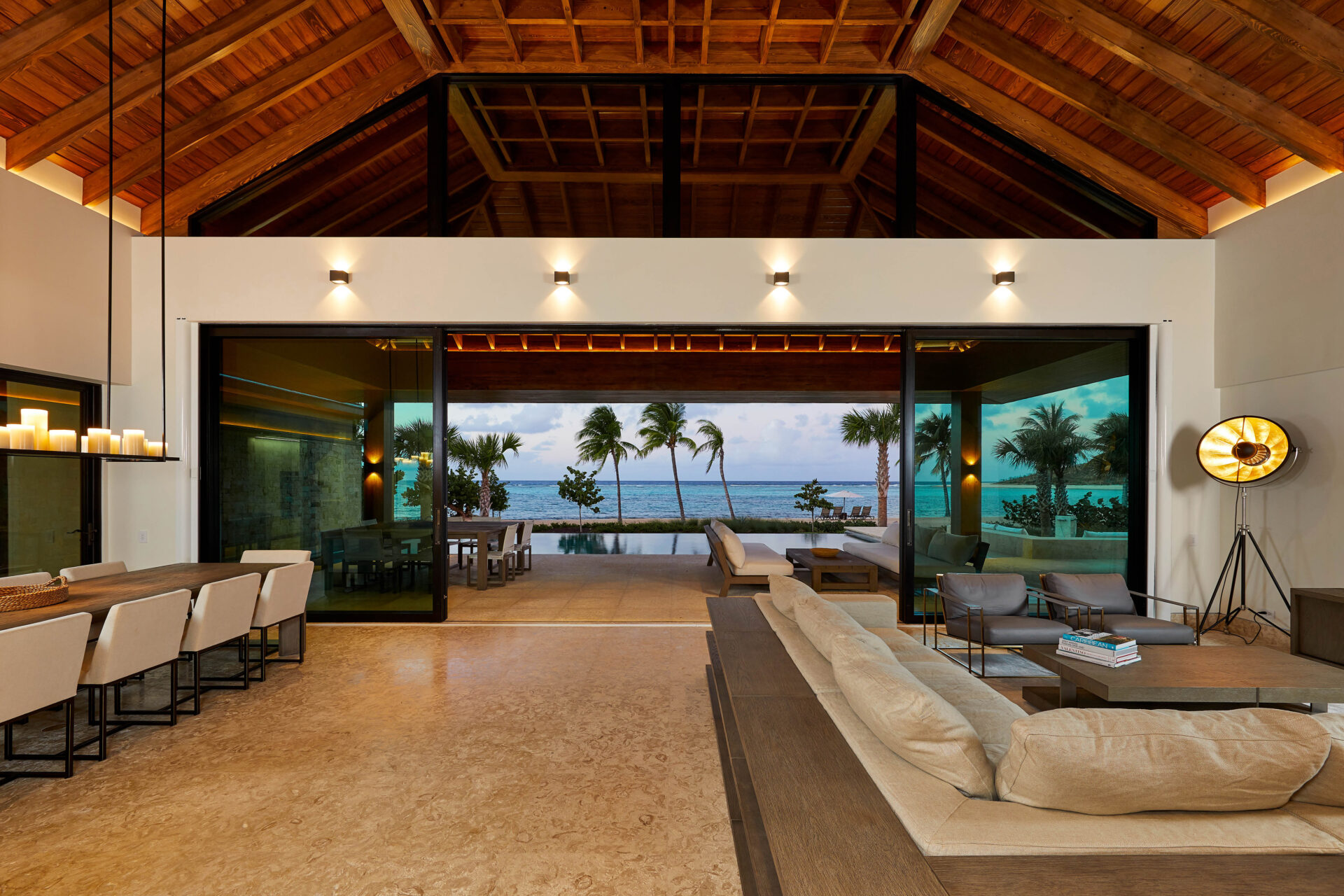 A spacious open-concept living room with high wooden ceilings opens up to a patio with ocean views in this stunning coastal rental. The modern interior features a large sectional sofa, a dining area with high-back chairs, and a kitchen island with bar stools. Palm trees and an infinity pool are visible outside.