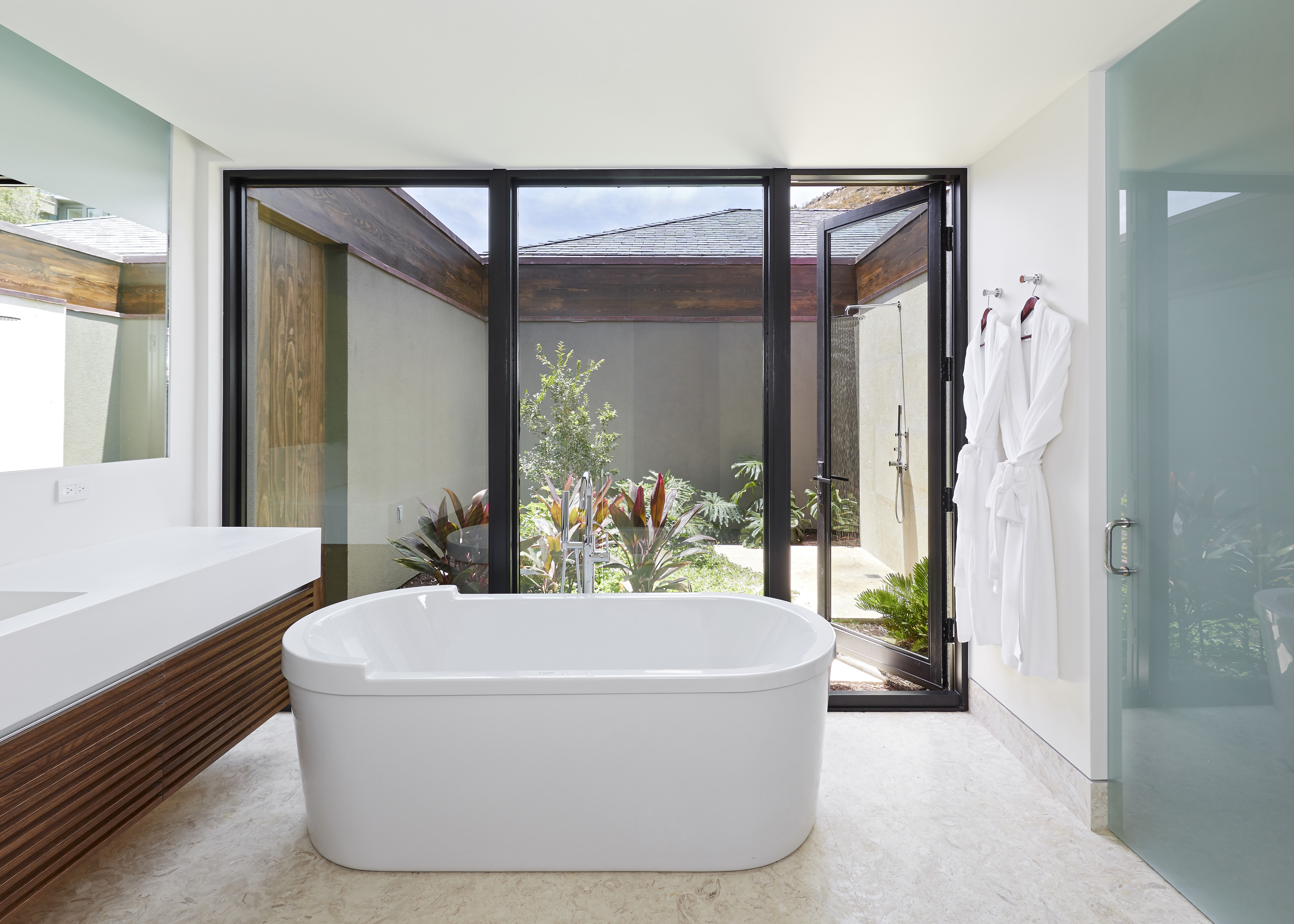 Modern bathroom with a freestanding bathtub positioned against a floor-to-ceiling window and sliding glass door, revealing an outdoor garden. Robes hang on the far wall, while a sleek vanity with a wooden finish and a large mirror grace the left side, adding to this beach house sanctuary.