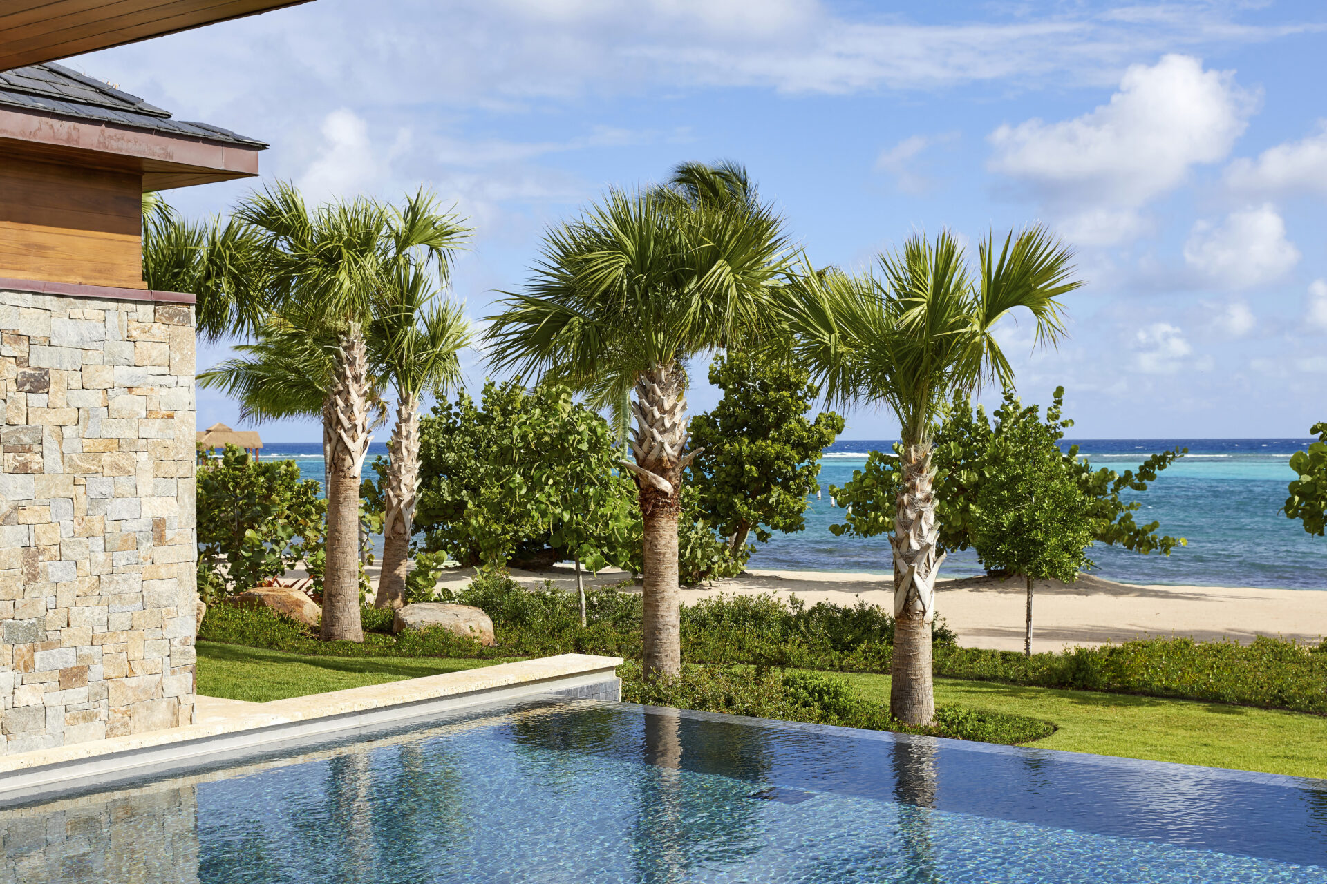 A luxurious poolside view features palm trees and a sandy beach against the backdrop of a clear, blue ocean and a partly cloudy sky. The edge of a stone beach house is visible on the left, adding to the serene and tropical ambiance.