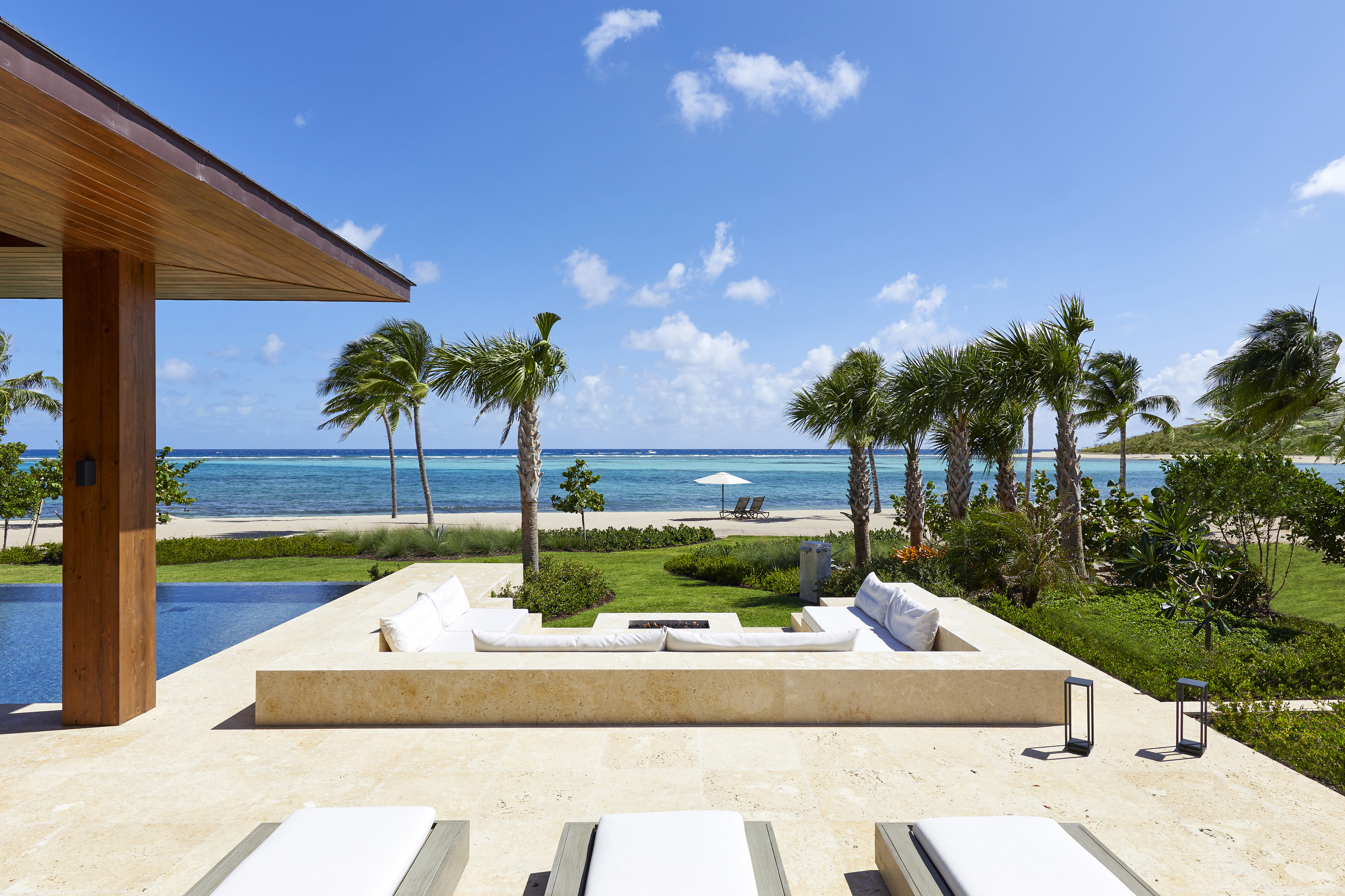 Luxury beachfront patio with white cushioned seating and lounge chairs, overlooking a pool, palm trees, and a turquoise ocean under a clear blue sky. This vacation rental features a shaded patio cover, lanterns, and an umbrella-shaded table, offering a serene and inviting atmosphere.