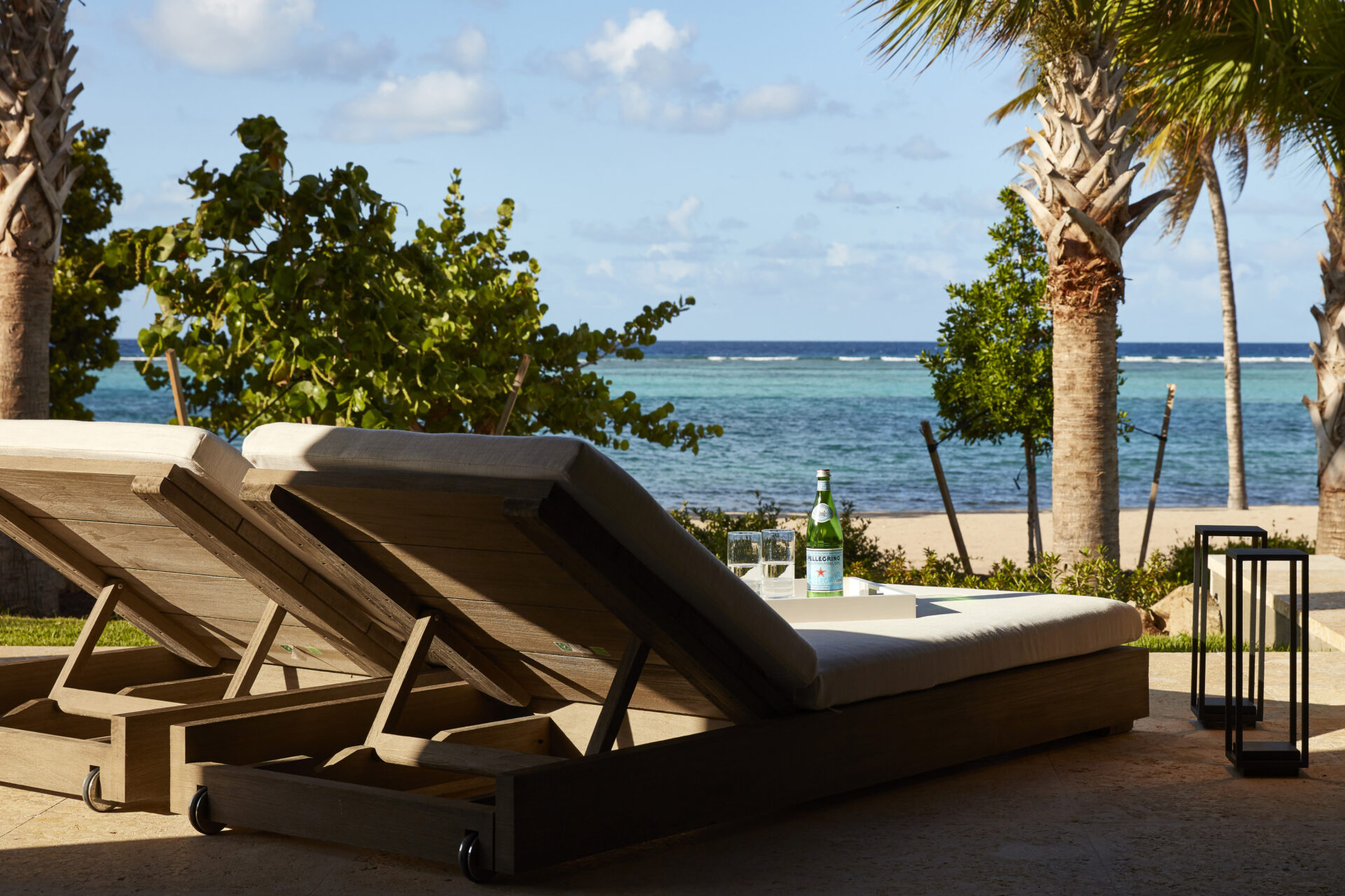 Two wooden lounge chairs with cushions are positioned under palm trees, facing a tranquil beach and the ocean. A small table between them holds bottles of water and glasses, suggesting a relaxing setup. The sky is partly cloudy, adding to the serene atmosphere near the beach house.