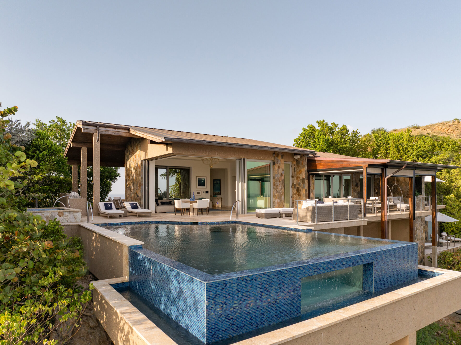 A modern, luxurious house known as Wild Side Villa features large glass doors and stone walls, complete with a spacious pool area. The infinite pool, tiled in blue, extends over the edge. Surrounded by lush greenery, this serene scene offers an elevated view of the landscape.