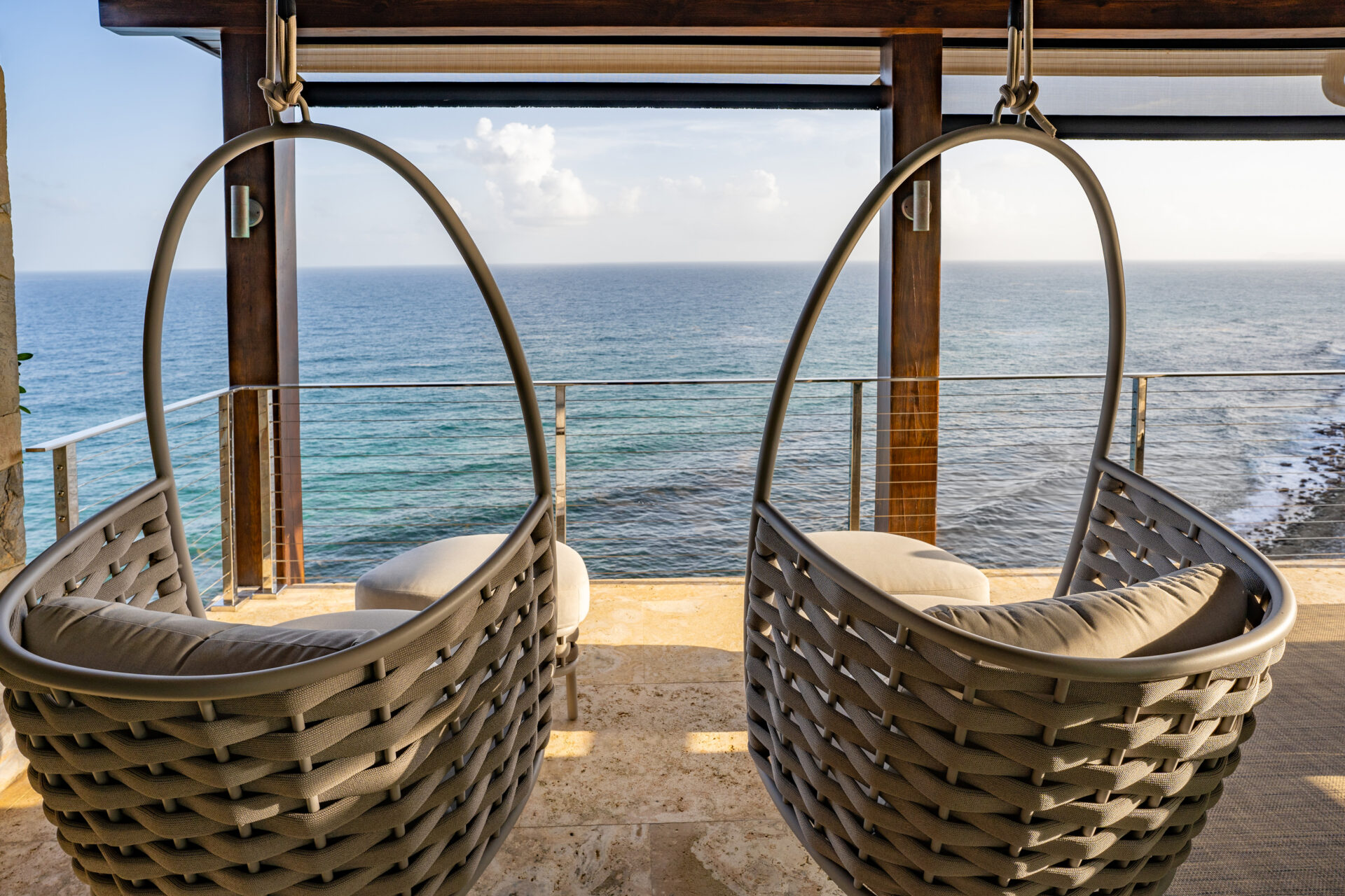 Two hanging wicker chairs with cushions are positioned on a balcony at Wild Side Villa, offering a direct view of the ocean. The sky is partly cloudy, and the calm water stretches to the horizon. The stone-tiled balcony floor adds to the relaxed, inviting ambiance of these luxury villa rentals.