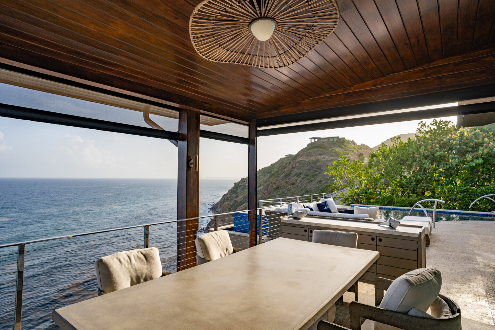 The Wild Side Villa boasts a covered outdoor dining area overlooking the ocean with a wooden ceiling and large modern light fixture. This villa rental features a long dining table with chairs, a lounge area with cushioned seating, and views of a forested hillside in the background.