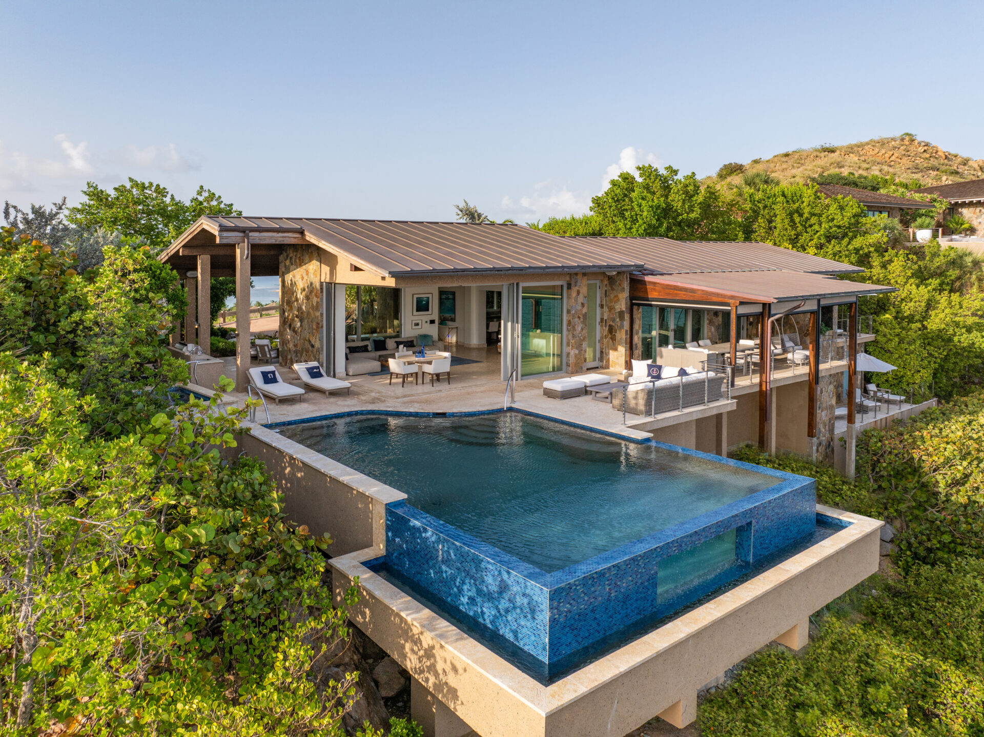 Wild Side Villa is a luxurious hillside retreat featuring a modern design with expansive glass windows and a large infinity pool. The house has multiple outdoor lounging areas and offers sweeping views over lush greenery and the ocean in the distance.