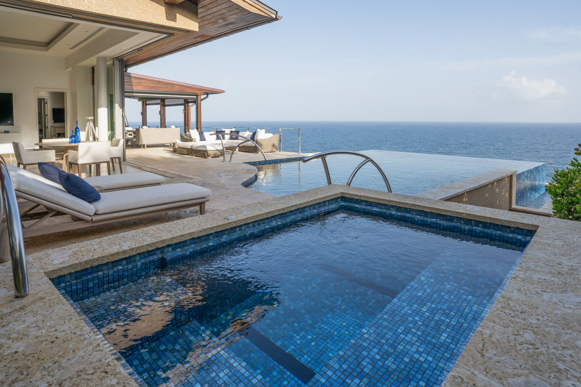 A luxurious outdoor pool area at Wild Side Villa features an infinity pool overlooking the ocean. The space includes sun loungers with cushions, a shaded lounging area with seating, and modern, elegant architecture. The view extends to a clear blue sky and calm sea in the background.