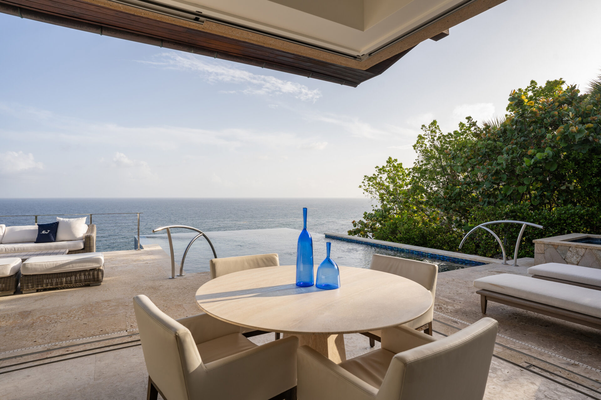 A serene outdoor patio at Wild Side Villa overlooks the ocean, featuring a round table with four chairs and three blue glass bottles. The space includes luxurious lounge chairs, an infinity pool, and lush greenery on the side. The sky is clear and sunny.