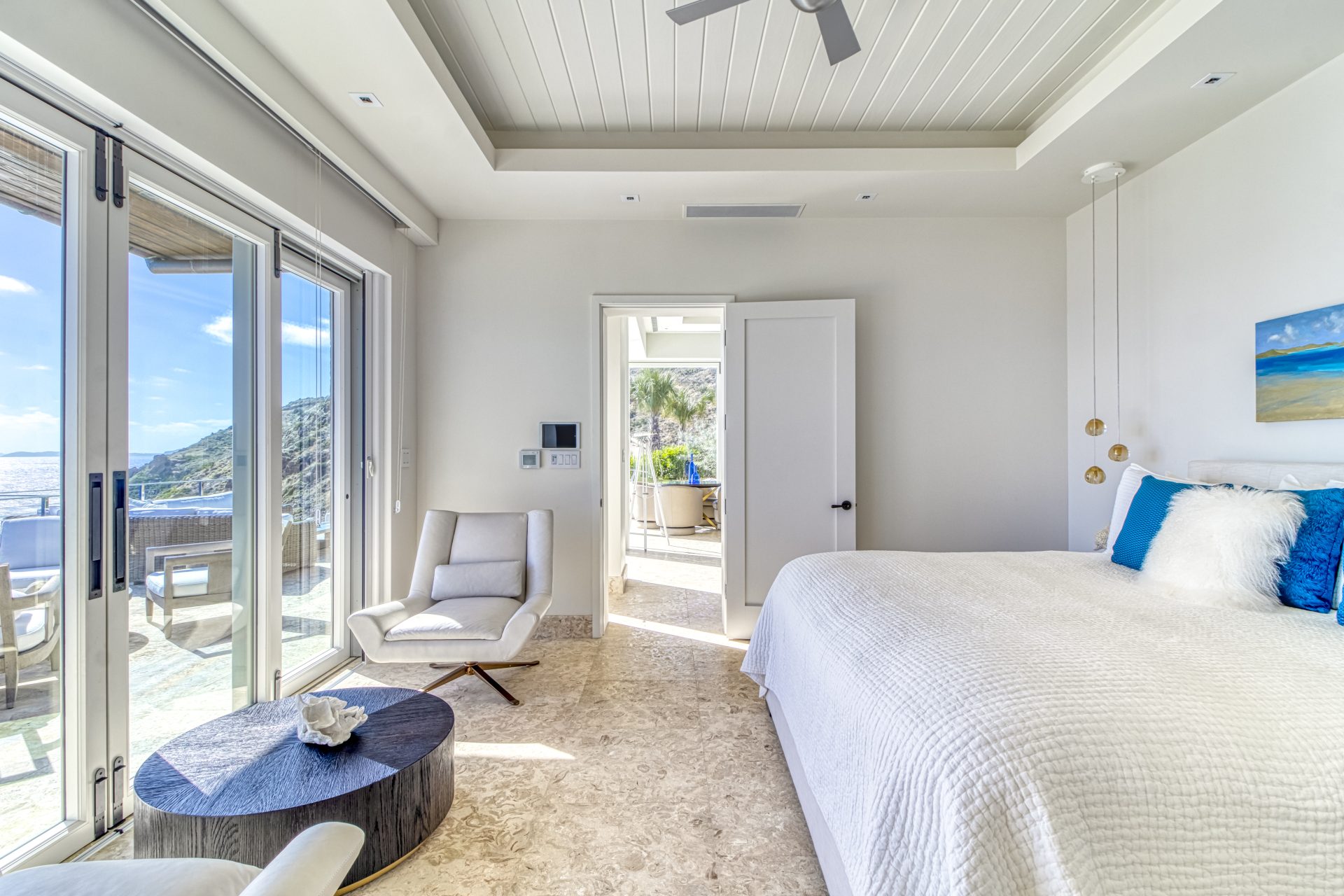 A bright, modern bedroom at Wild Side Villa boasts a white bed and blue accents. The room features a cozy armchair, round coffee table, and large glass doors leading to a balcony with an ocean view. A doorway at the far end opens to a sunlit patio.