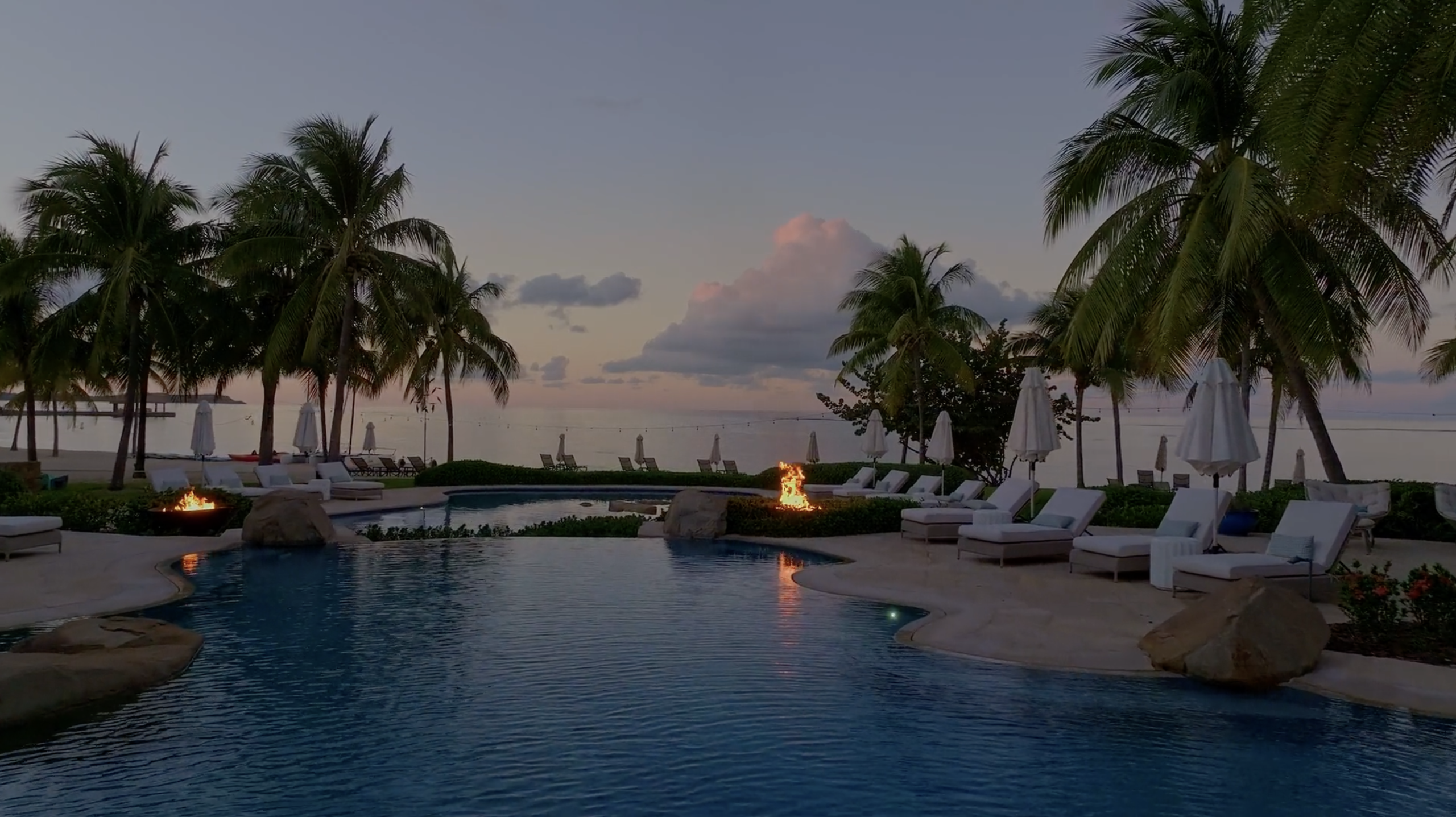 A tranquil beach resort at dusk, featuring a luxurious pool surrounded by lounge chairs and palm trees. A pavilion houses a charming bar near the beach, while flaming torches light the path to the calm ocean and vibrant sunset sky in the background.