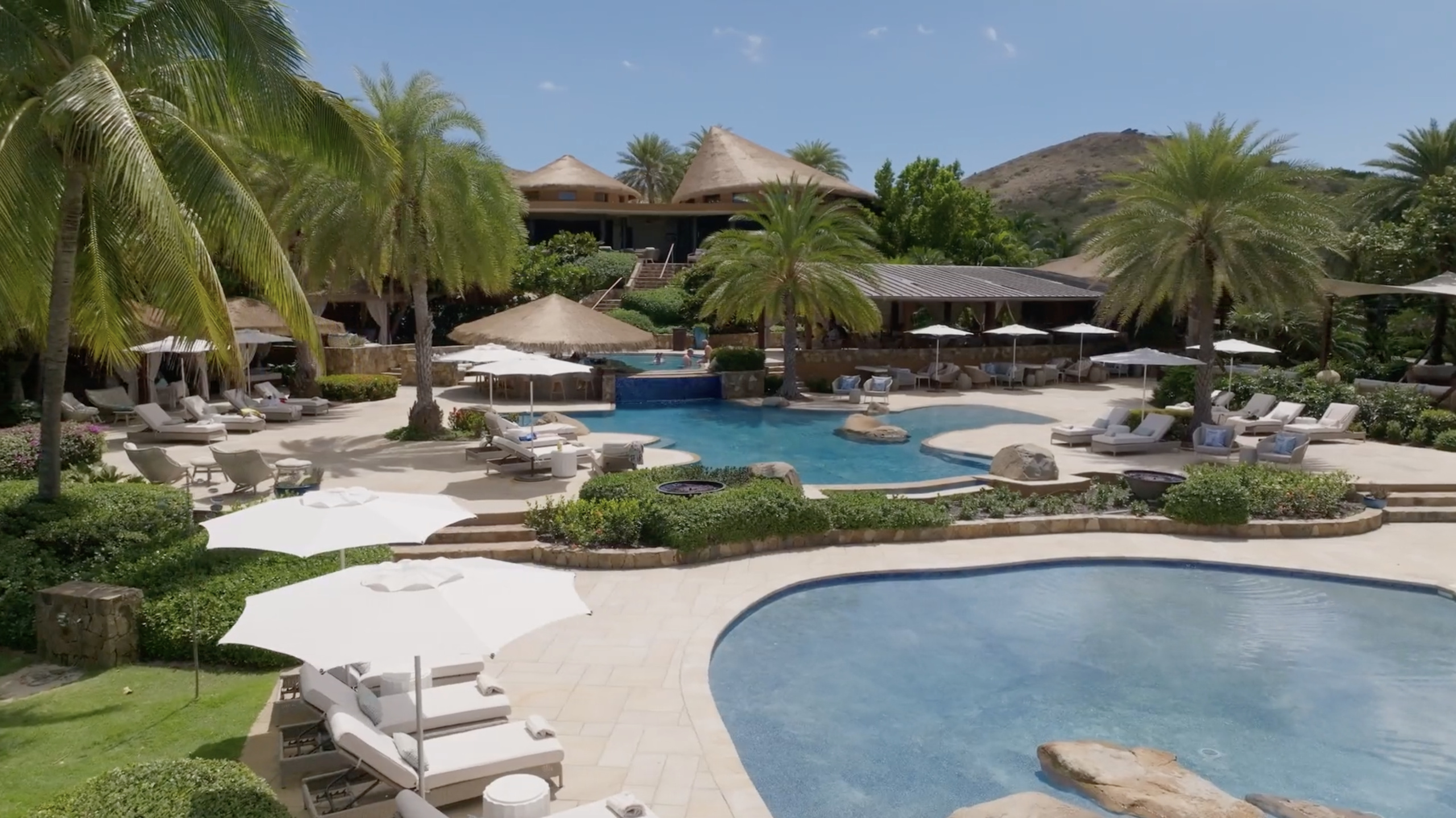 A luxurious tropical resort features multiple swimming pools, sunloungers with white umbrellas, and lush palm trees. In the background, there are thatched-roof buildings set against a backdrop of green hills and clear blue skies.