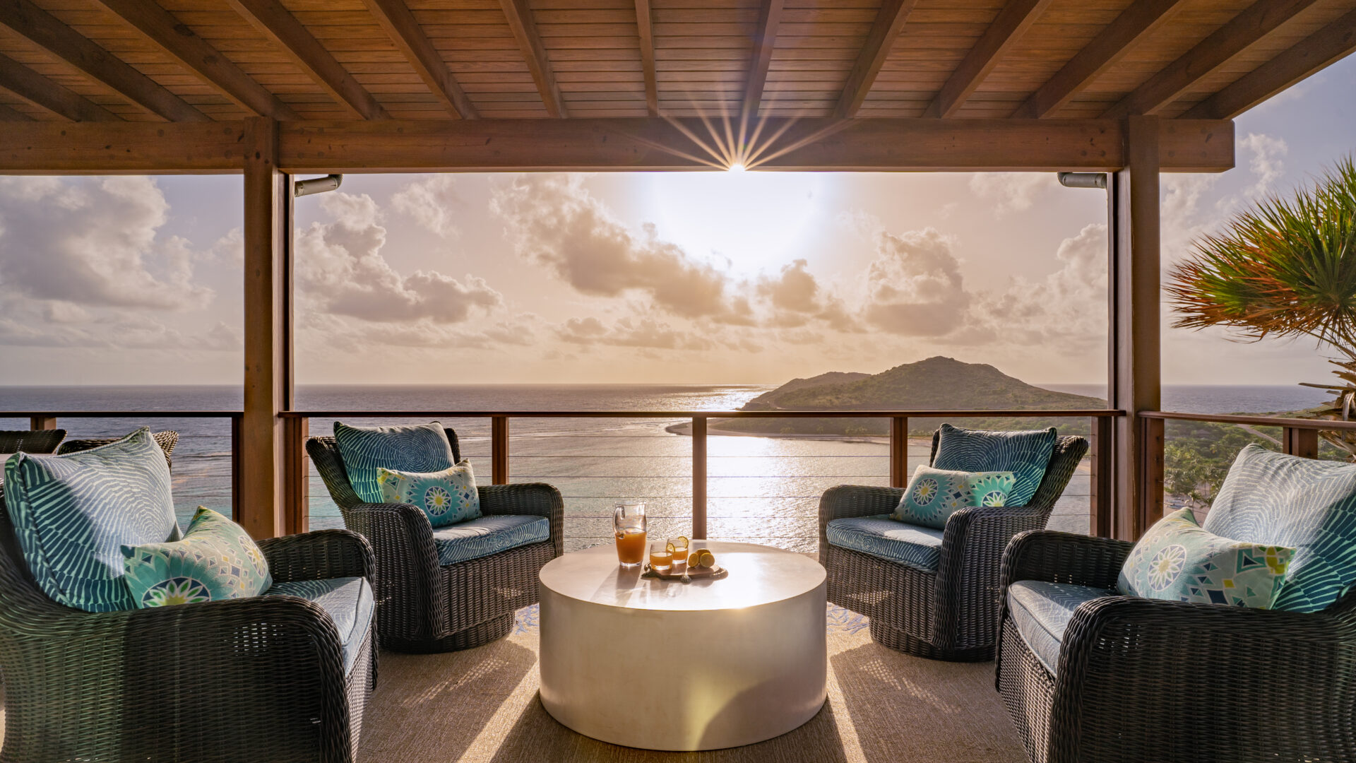 An outdoor seating area at Poseidon's Perch Villa features four cushioned wicker chairs surrounding a round table with glasses of beverages. The area overlooks the ocean, offering a scenic view of an island and a sun setting behind clouds. The space is enhanced by wooden roofing and railing.