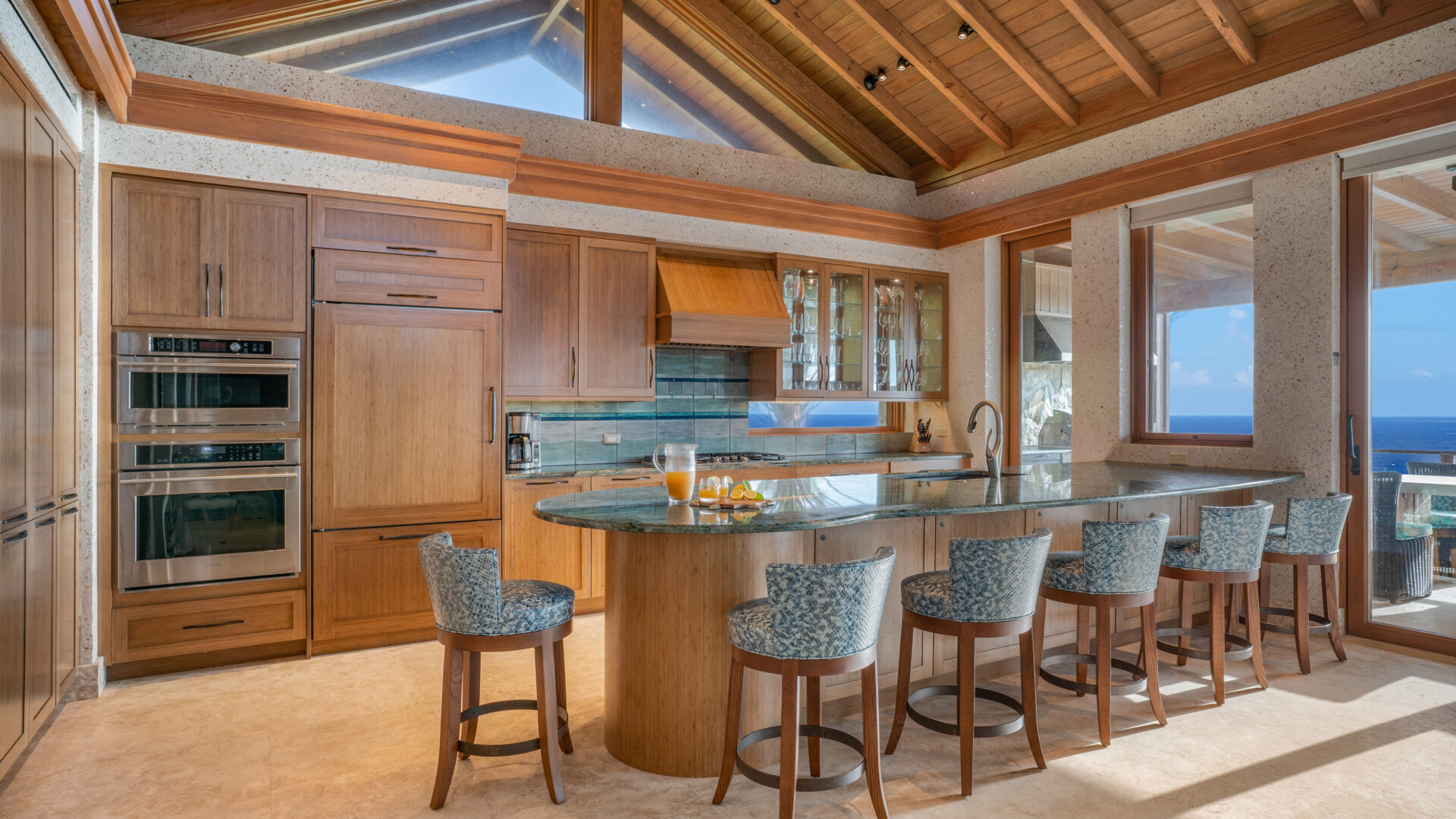 A spacious kitchen with wooden cabinetry and high vaulted ceilings. It features a large kitchen island with blue patterned bar stools, built-in oven, and a scenic ocean view visible through large windows. The ceiling has exposed wooden beams and natural light floods in, perfect for Villa Poseidon's Perch.