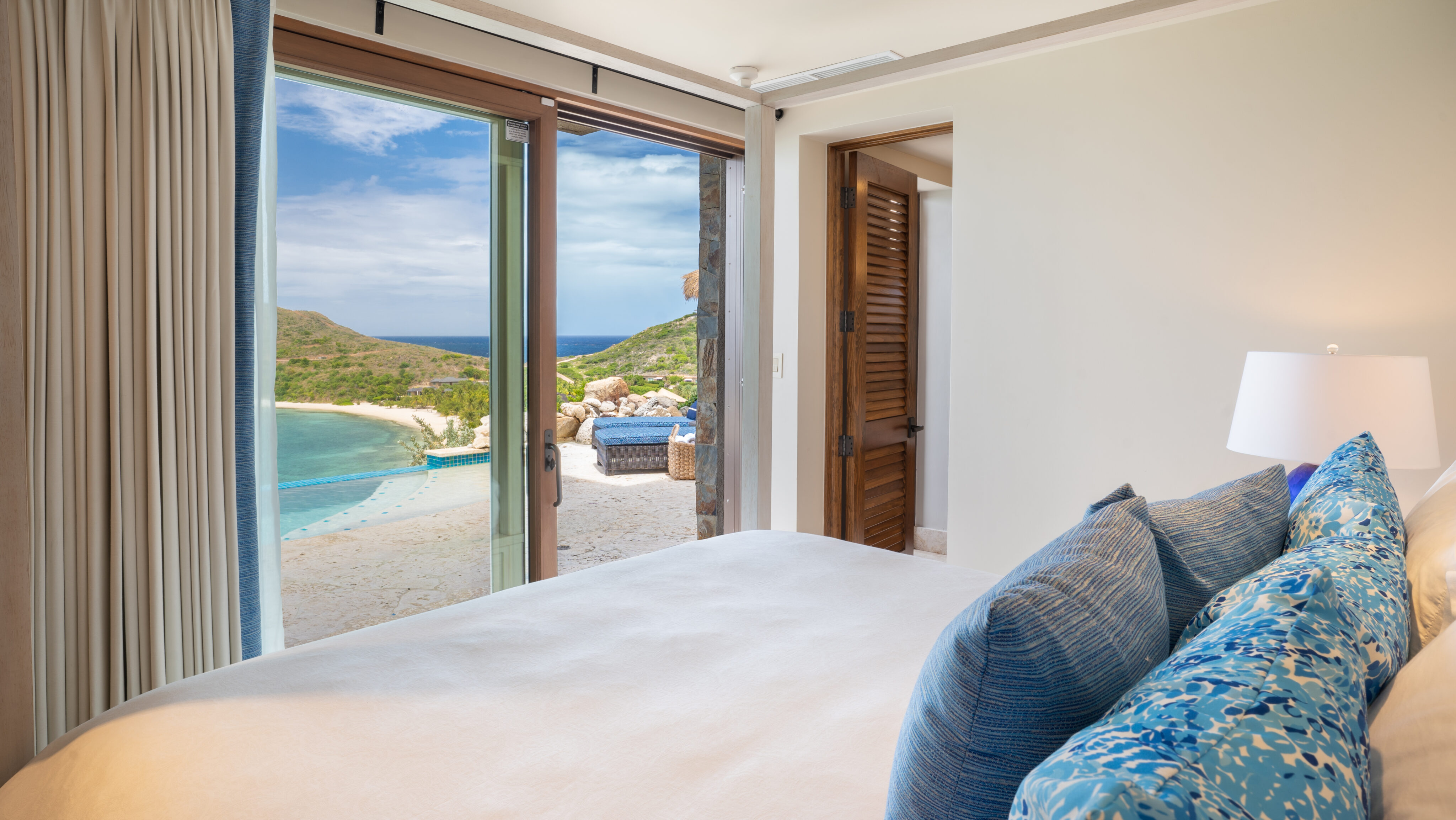 A serene bedroom at Villa Poseidon's Perch with blue and white decor features an open sliding glass door leading to a private beach with a calm, clear ocean and lush, green hills in the background. A wooden door stands ajar, revealing the cozy, inviting nature of the room.