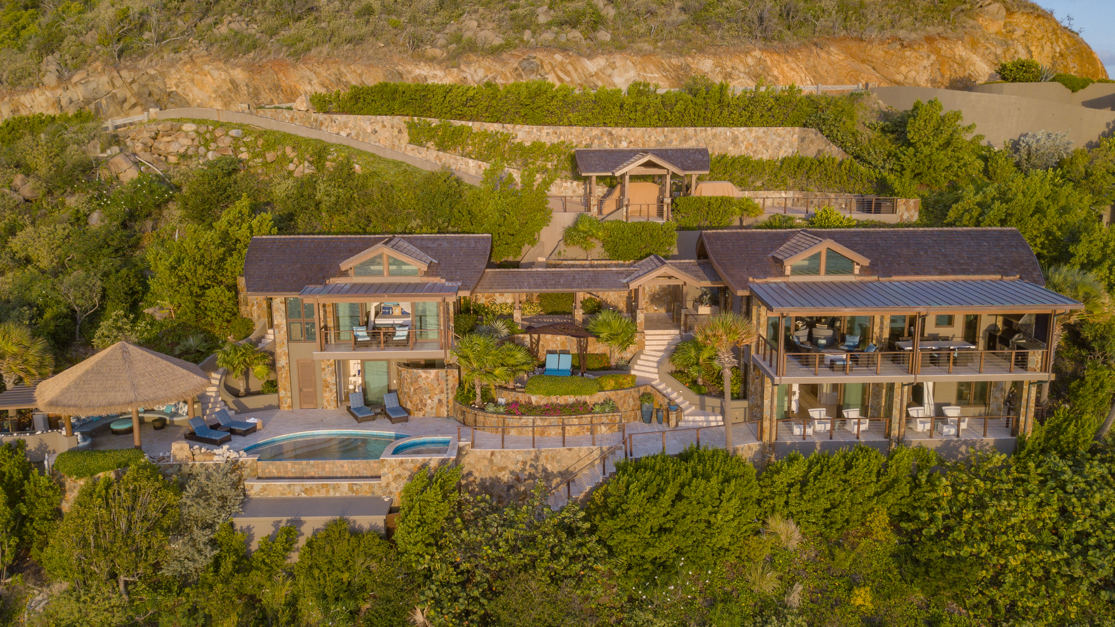 Aerial view of Poseidon's Perch Villa, a luxurious hillside estate with multiple buildings, an infinity pool, and lush greenery. The property includes a main house, guest houses, outdoor seating areas, and scenic landscape featuring stone walls and terraces.