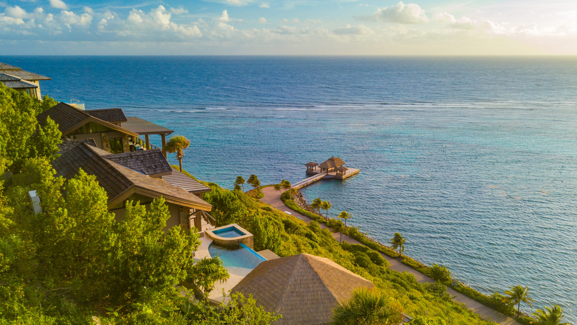 A scenic view of a coastline with several houses nestled on a hillside. Below, Poseidon's Perch Villa offers luxury accommodation with a pool surrounded by lush greenery and a path leading to a small pier stretching into the calm, blue ocean. The horizon is clear with a few scattered clouds in the sky.