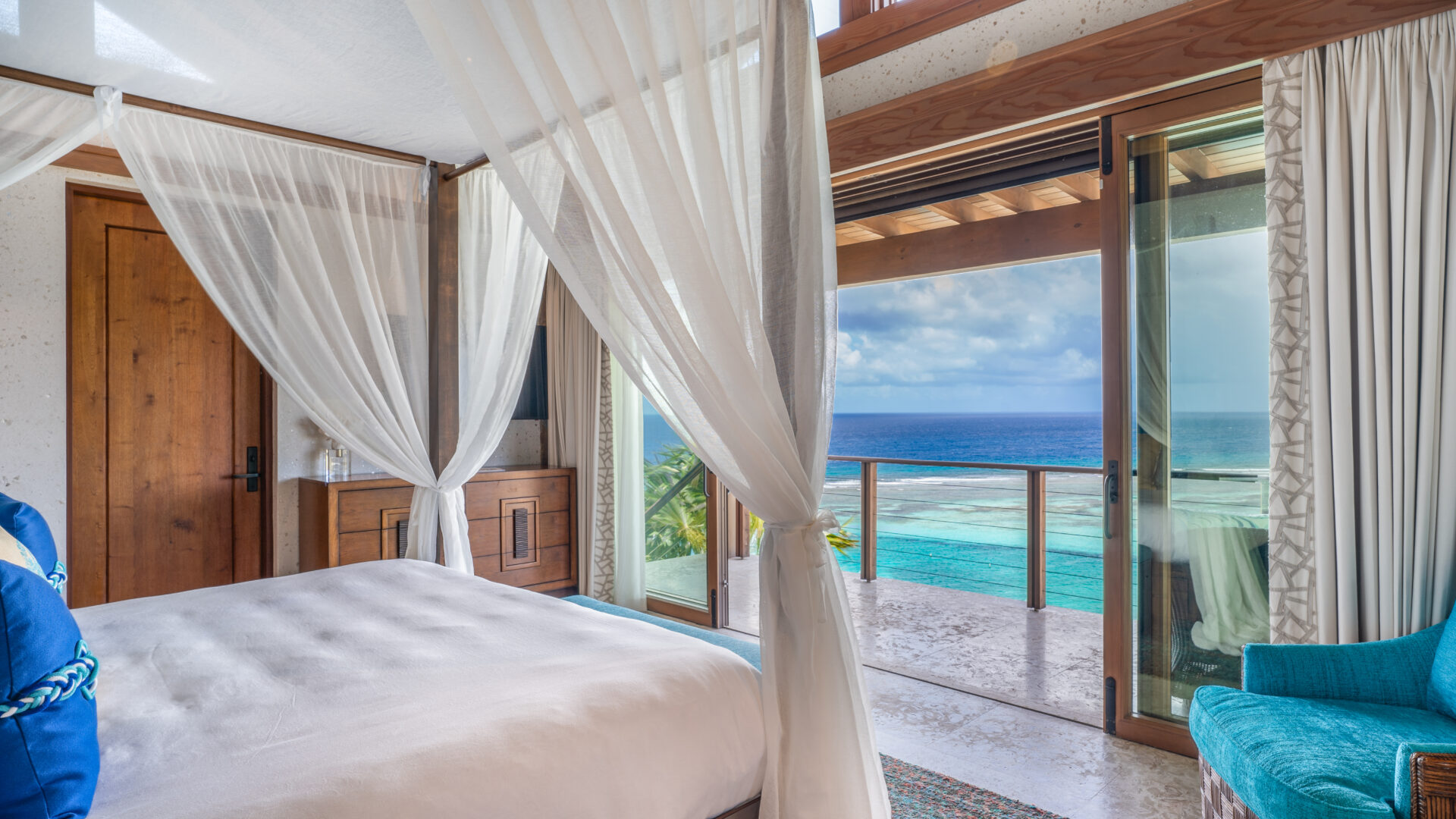 A serene bedroom at Poseidon's Perch Villa features white curtains and a canopy bed, opening to a balcony with stunning ocean views. Wooden furniture and a turquoise armchair enhance the room's coastal feel, while sliding glass doors provide plenty of natural light in this luxury accommodation.