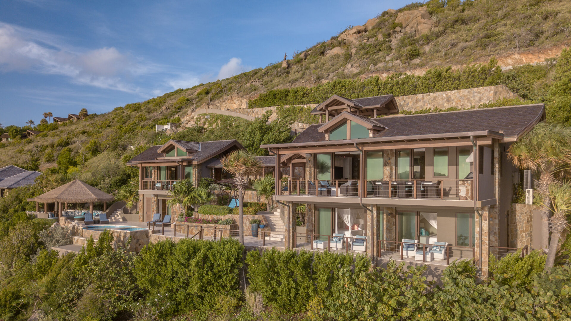 Poseidon's Perch Villa, a luxurious hillside complex, boasts two multi-story houses with balconies and expansive windows. A swimming pool and thatched gazebo lie to the left, surrounded by lush greenery and vibrant landscaping under a clear blue sky.