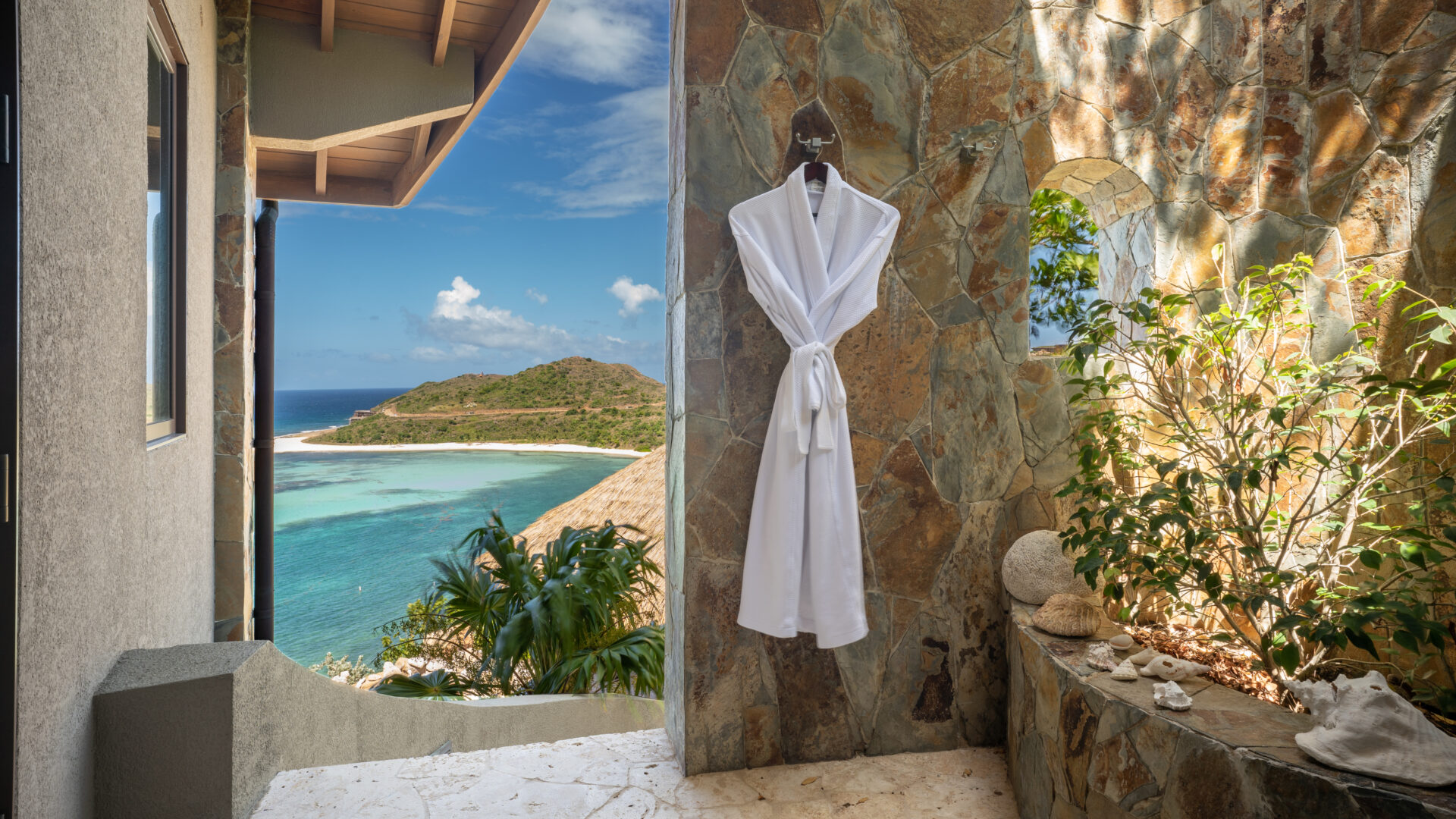 A white bathrobe hangs on a stone wall in an open-air bathroom at Poseidon's Perch Villa, boasting a stunning view of an island and the turquoise ocean. Tropical plants adorn the space, while a wooden roof extends from the building, epitomizing luxury villa rental with oceanfront elegance.