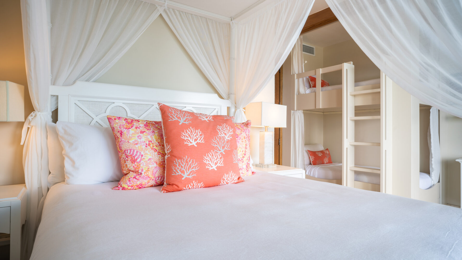 A bright and airy bedroom in Poseidon's Perch Villa boasts a white canopy bed adorned with coral-patterned pillows. The room also features a set of white bunk beds against the wall, each with its own coral-themed pillow. Light filters softly through the sheer canopy curtains, exuding the feel of a luxury villa rental.