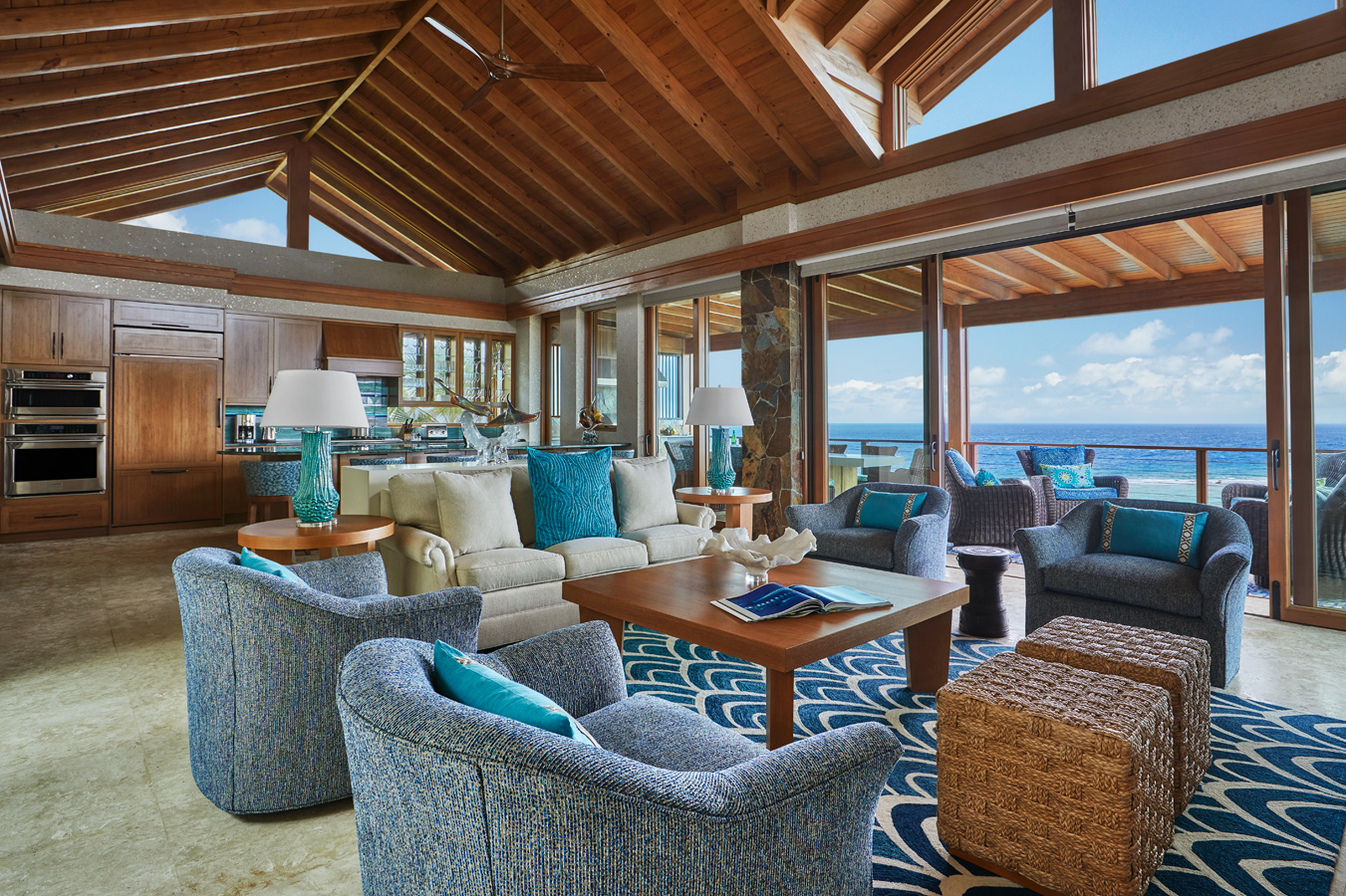 Experience the luxurious open-plan living area of Poseidon's Perch Villa, featuring a vaulted wooden ceiling and large glass doors that offer a stunning ocean view. The room is furnished with blue and brown chairs, a wooden coffee table, decorative cushions, and modern kitchen cabinets in the background.