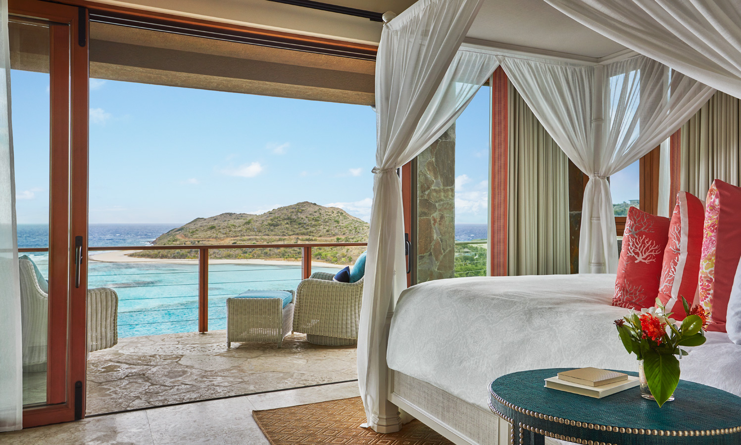 A luxurious bedroom at Poseidon's Perch Villa features a four-poster bed adorned with white drapes and pink pillows. The room has sliding glass doors that open to a balcony overlooking a scenic ocean view and a hilly landscape. In the foreground, there is a round table holding books and a flower vase.