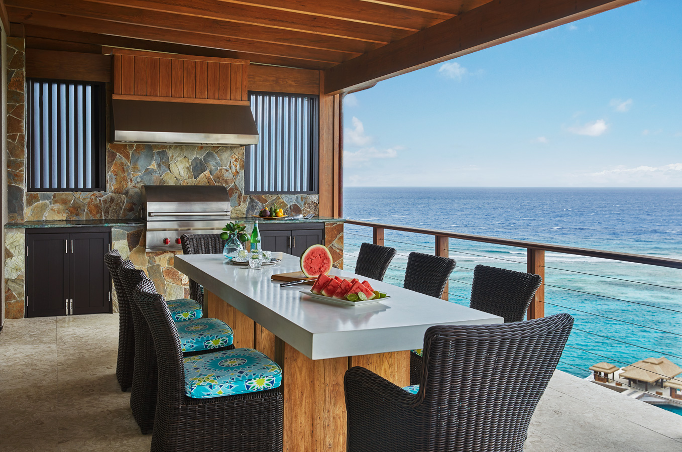 At Poseidon's Perch Villa, the outdoor kitchen and dining area offer a breathtaking ocean view. Features include a stone grill, countertop with a sliced watermelon, and a long dining table with black wicker chairs adorned with blue floral cushions. The space is enhanced by a wooden ceiling and railing.