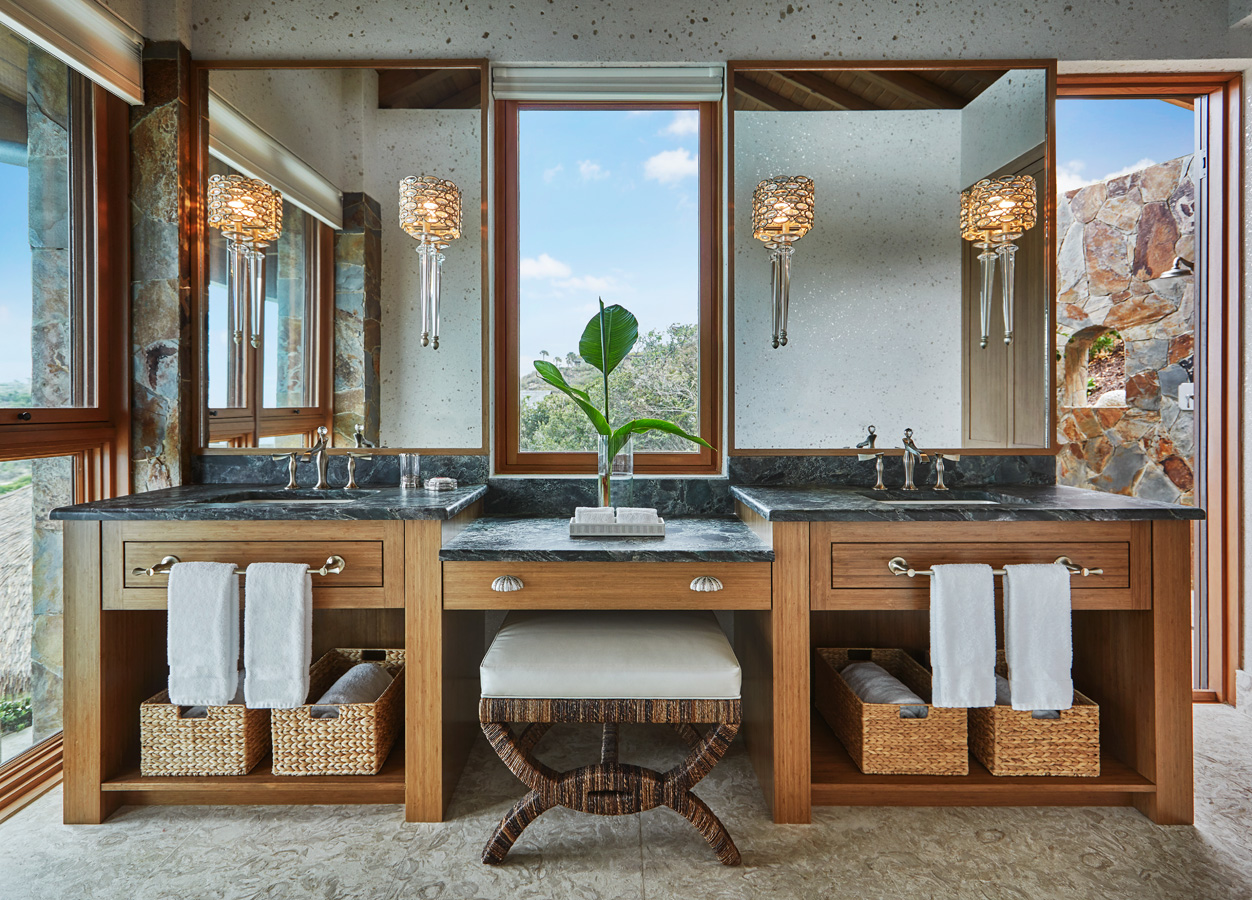 A luxurious bathroom in Poseidon's Perch Villa features two dark marble sinks with wooden cabinets, each adorned with a wicker basket holding a white towel. A large mirror sits above each sink, flanked by elegant wall sconces. A window with a view is centered between the mirrors.