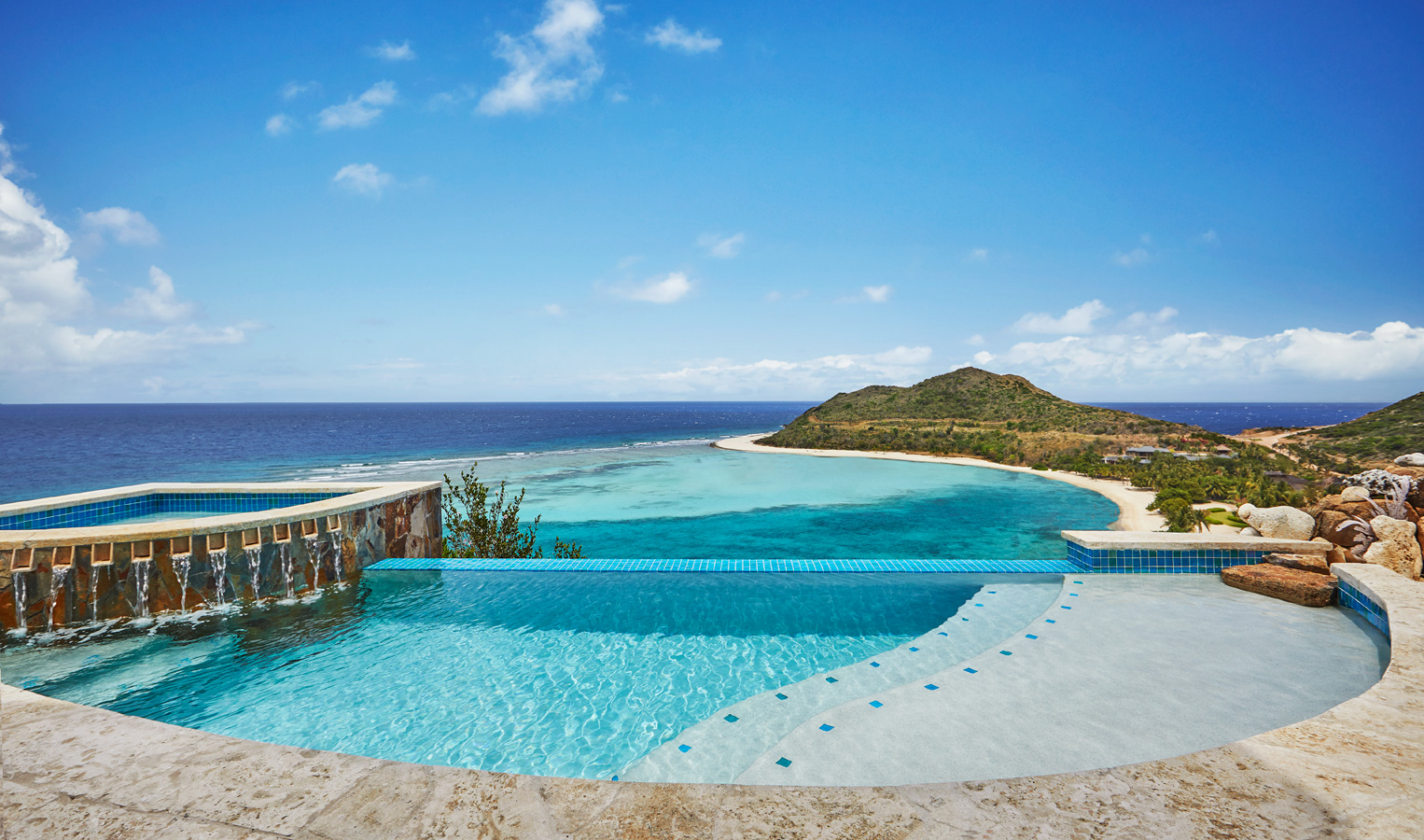 A luxurious infinity pool with a waterfall edge, part of the elegant Poseidon's Perch villa, overlooks a pristine beach and a serene ocean bay. The scene includes a lush, hilly island under a bright blue sky with scattered clouds, exuding relaxation and natural beauty.