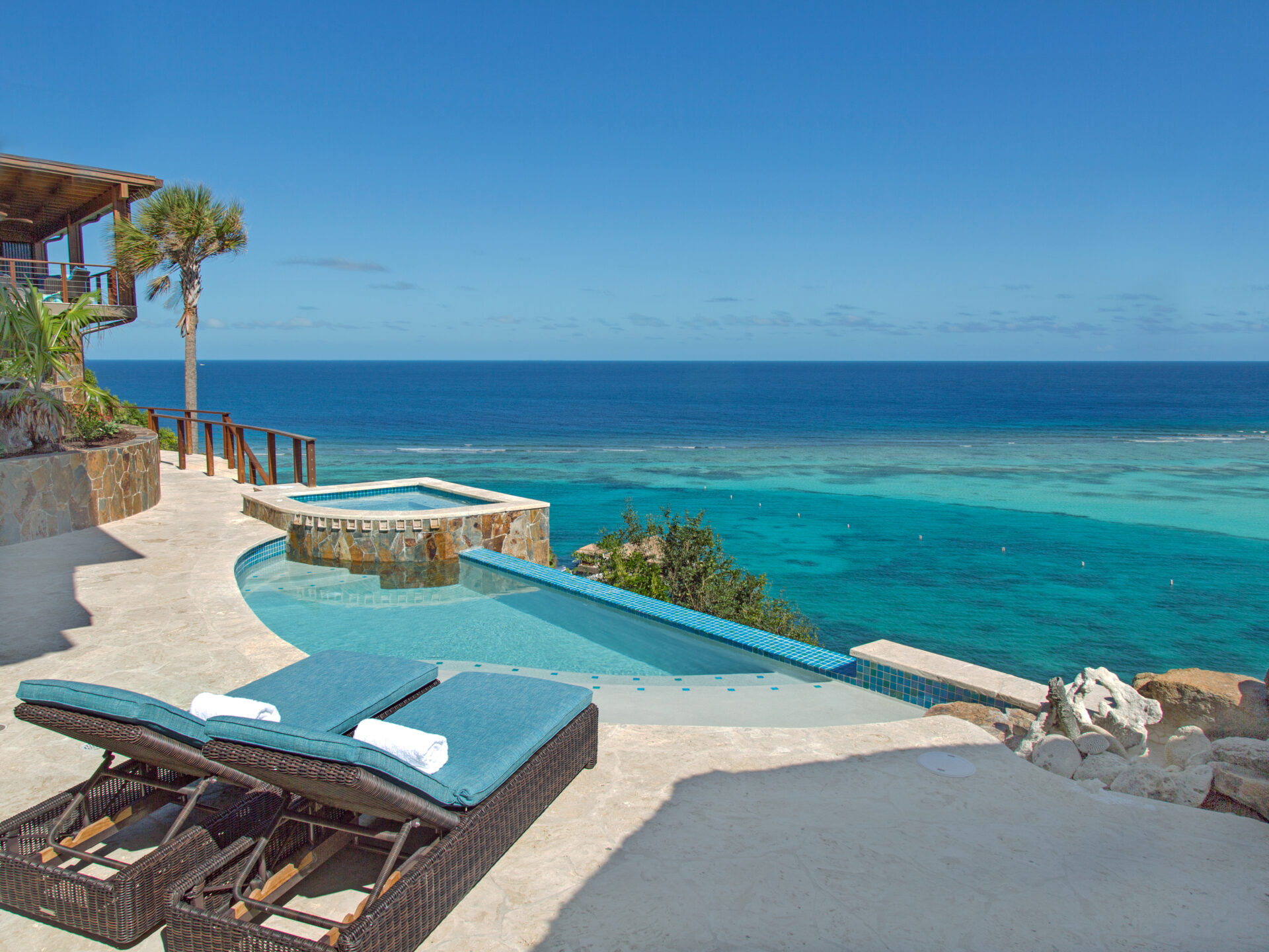 A scenic coastal view at Poseidon's Perch villa features an infinity pool overlooking a turquoise ocean. On the patio, two cushioned lounge chairs are positioned beside the pool, which is bordered by stone walls and tropical plants. The horizon meets a clear blue sky.