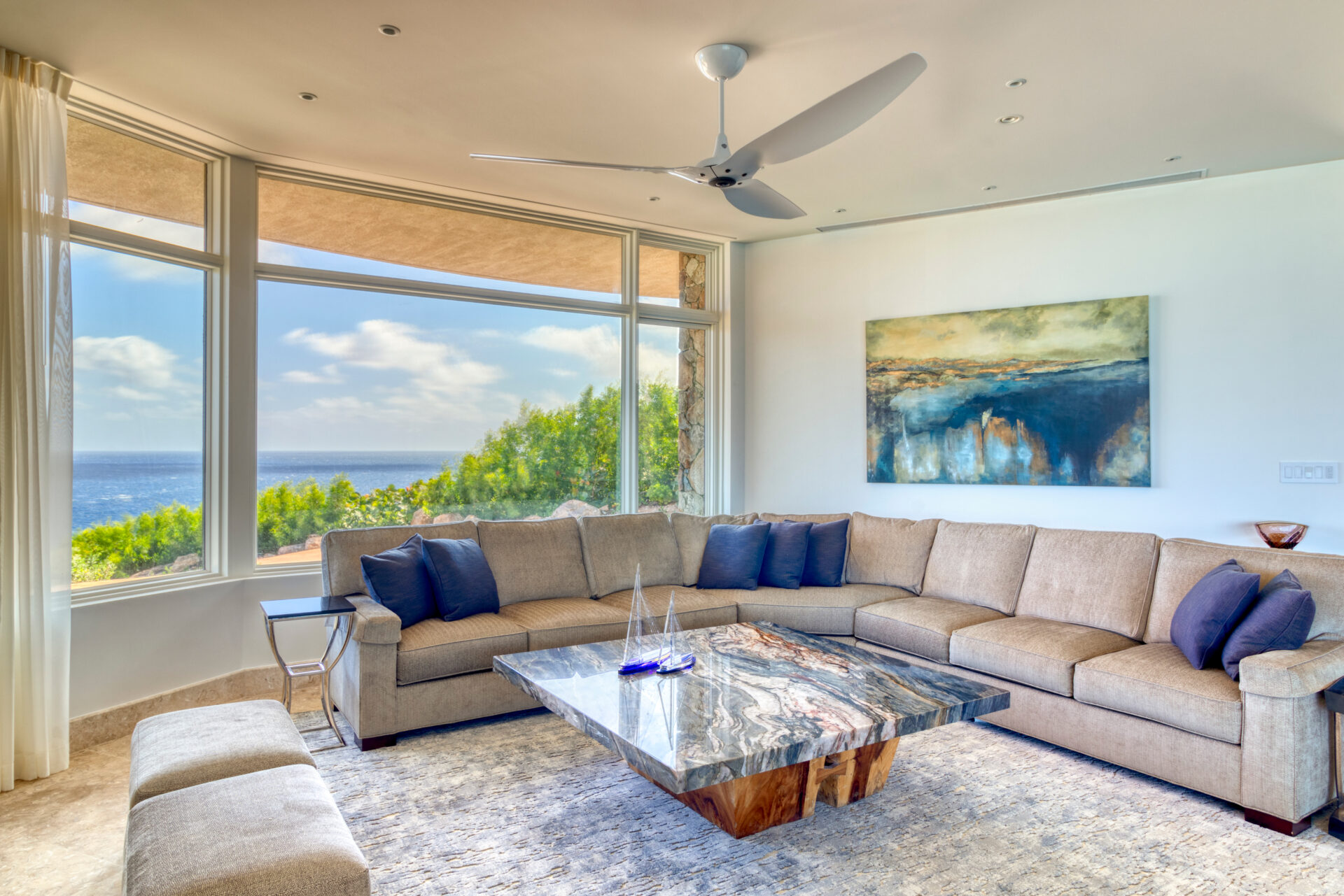 Nestled within Rainbow Villa, this bright and airy living room boasts large windows showcasing a scenic ocean view. The space features a beige sectional sofa with blue pillows, a unique marble coffee table, abstract art on the wall, a ceiling fan, and white curtains.