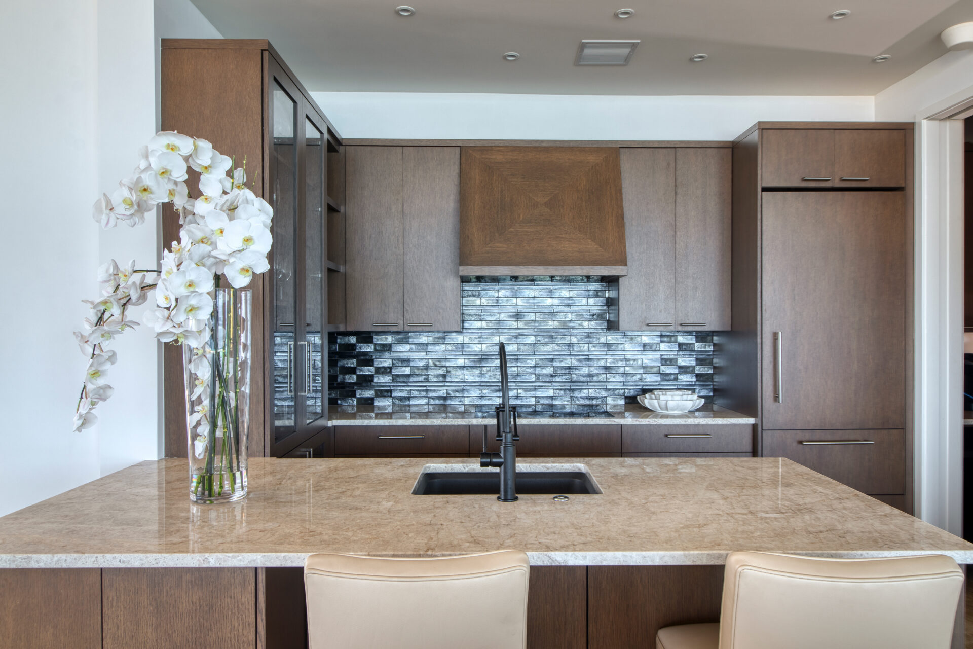 A modern kitchen at Rainbow Villa boasts wooden cabinets and a marble countertop island with a built-in sink and black faucet. A tall vase with white orchids graces the island. The backsplash features metallic tiles, complemented by beige bar stools.