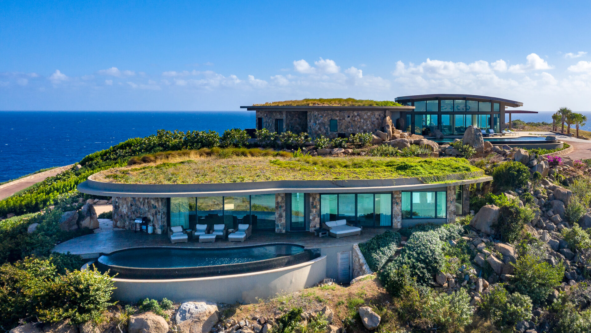 Rainbow Villa is a modern, luxurious cliffside retreat overlooking the ocean with expansive glass walls, multiple levels, and lush greenery on the roofs. The property features a spacious patio with seating and an infinity pool, surrounded by natural rock formations and vegetation.