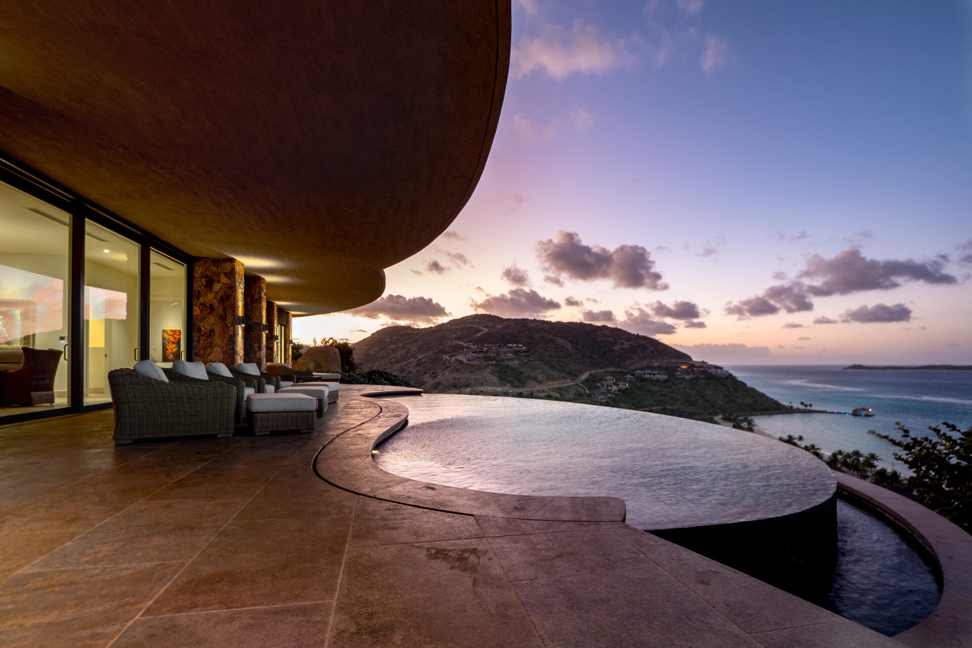 A modern patio at dusk with comfortable seating overlooks an infinity pool and a scenic view of the ocean and distant hills. At Rainbow Villa, the horizon shows a sky transitioning in hues of purple and orange as the sun sets.