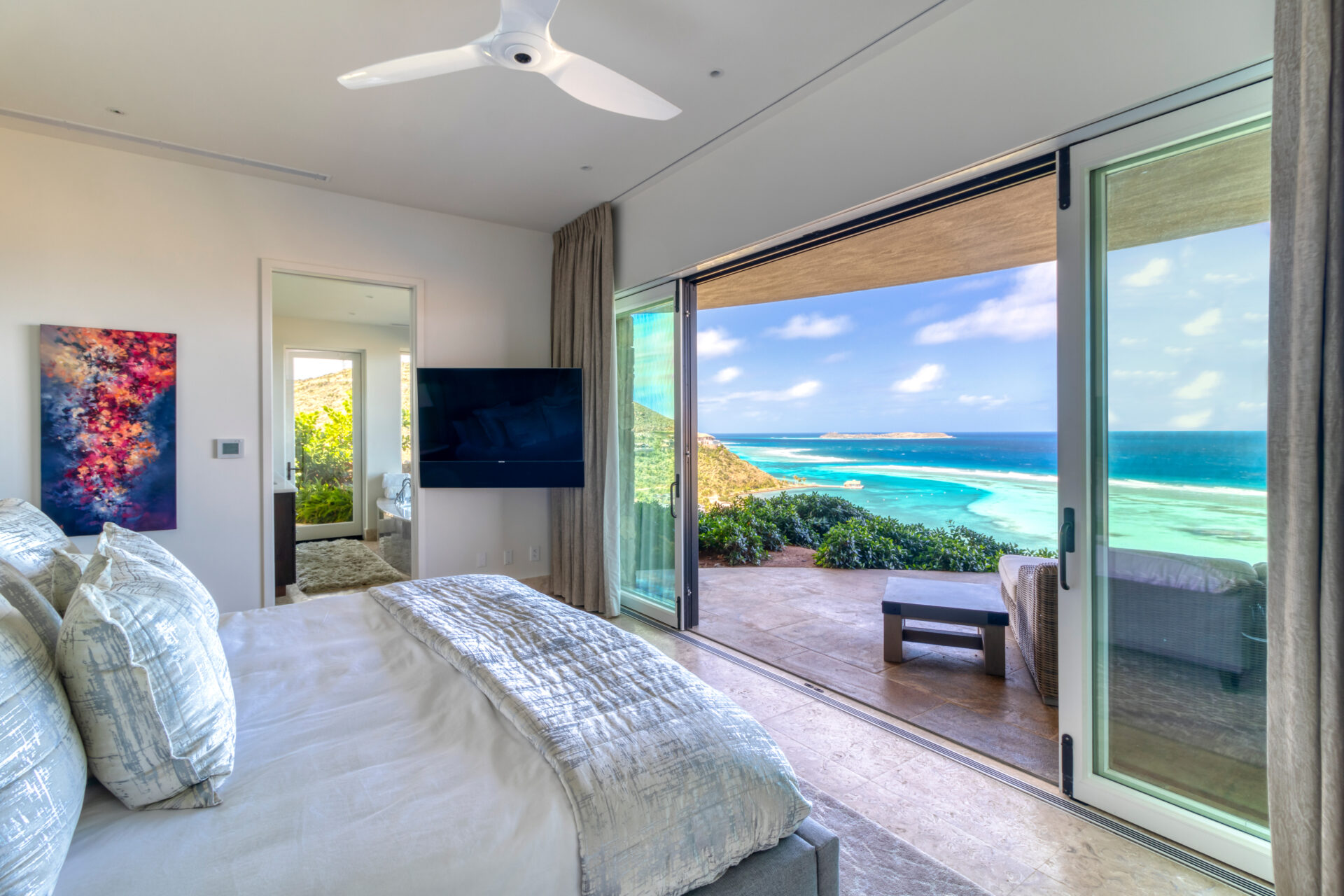 A modern bedroom in Rainbow Villa features a large bed, wall-mounted TV, and captivating artwork. Glass sliding doors open to a spacious balcony with seating, offering a stunning view of the ocean and coastline. A ceiling fan is mounted above, and soft natural light fills the room.