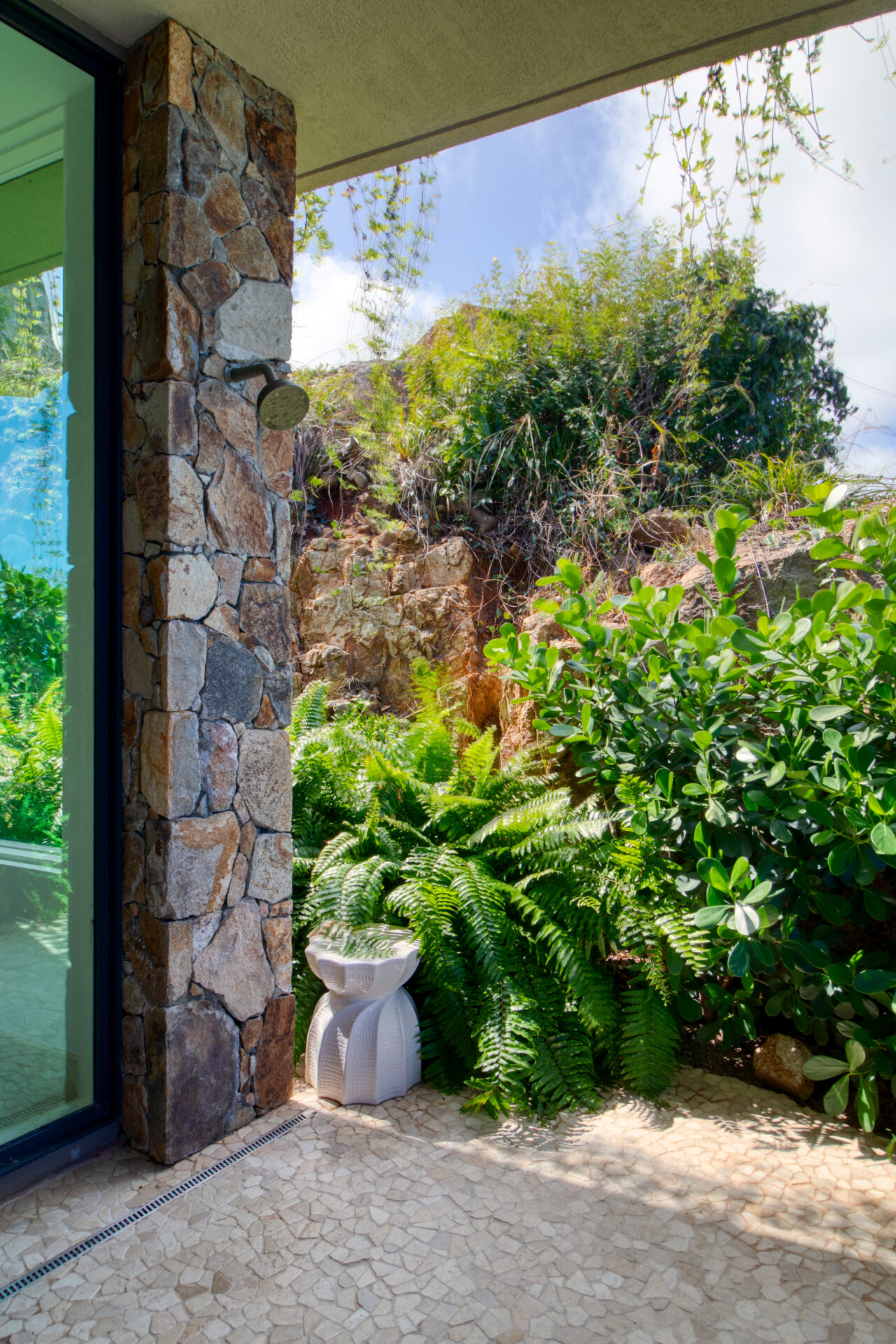 An outdoor shower area at Rainbow Villa is surrounded by lush greenery, including ferns and bushes, with a stone wall and a glass door leading to an adjacent room. The area is paved with textured tiles, and a decorative stool is placed near the shower.