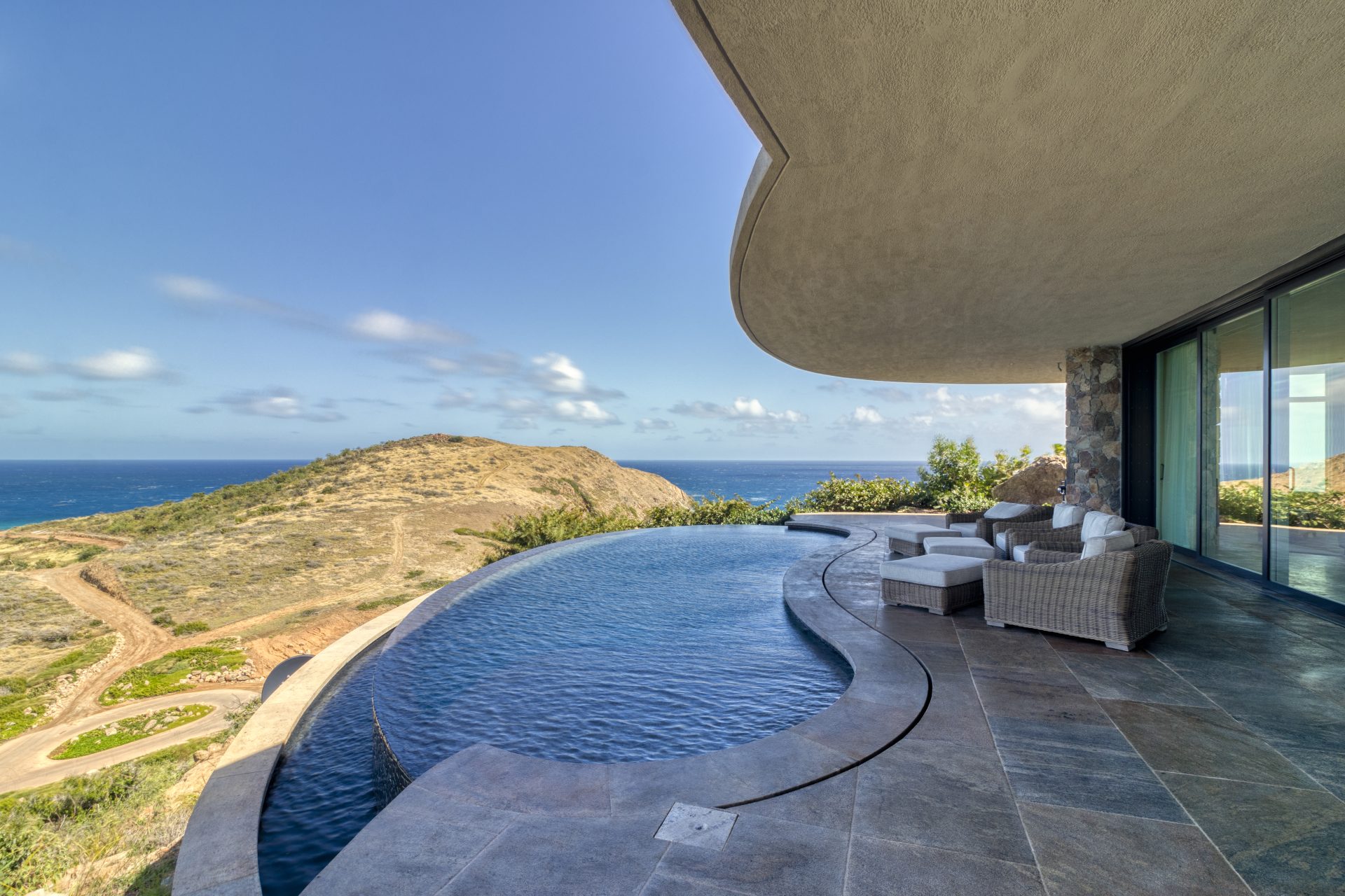 Rainbow Villa’s luxurious infinity pool seamlessly blends into the ocean view, with curved architecture creating shade over the patio. Plush outdoor furniture sits by the pool, and the landscape features rolling hills leading to the sea under a bright, partly cloudy sky.