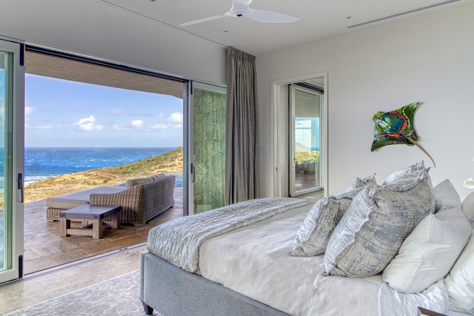 A bright bedroom in the Rainbow Villa features a large bed adorned with white and gray pillows and linens, complemented by a wall-mounted fish sculpture. Glass sliding doors open to a patio with outdoor furniture and a stunning view of the rugged coastline and ocean under a cloudy sky.