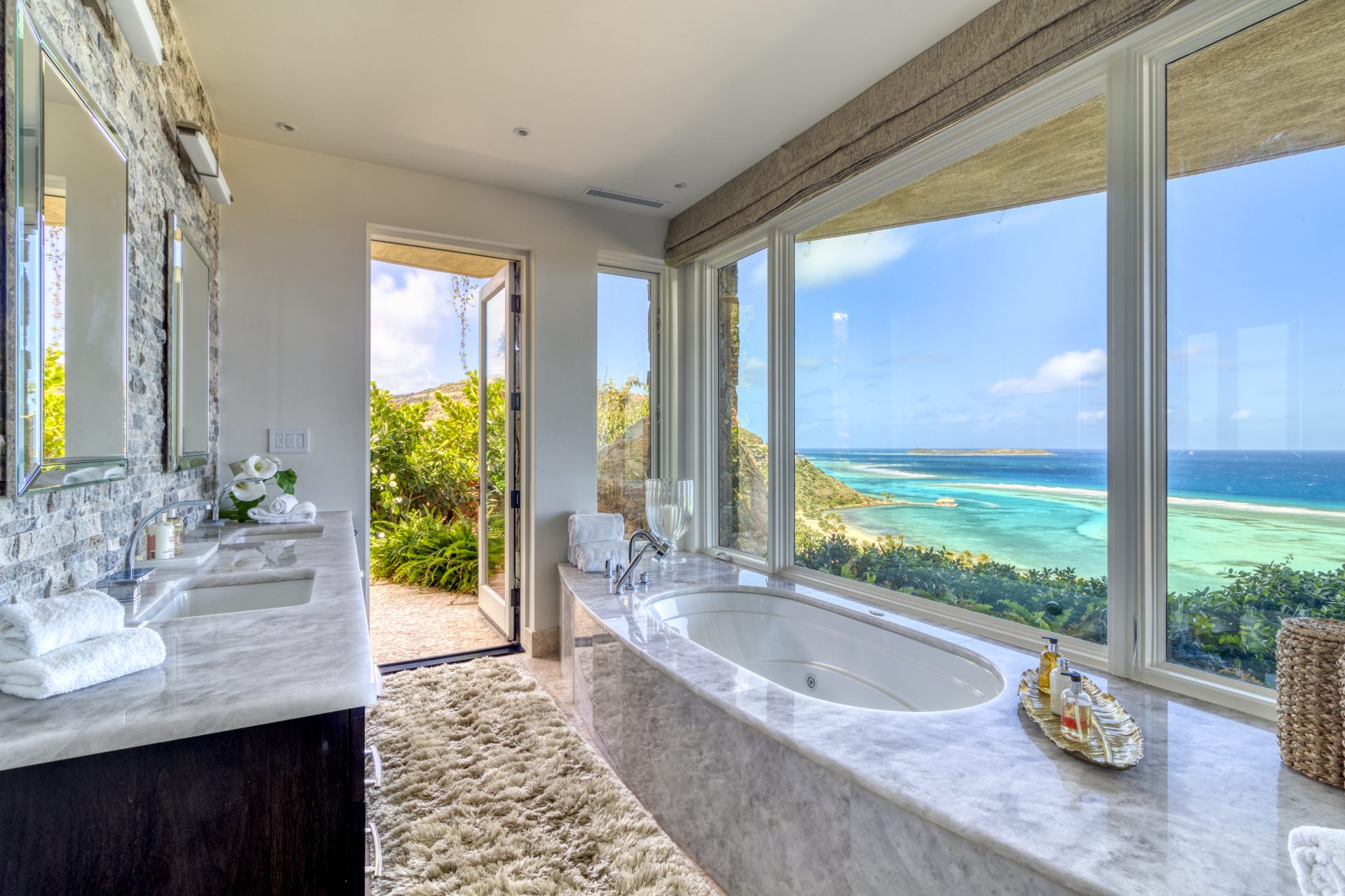 Luxurious bathroom at Rainbow Villa with large windows offering a stunning ocean view. Features a spacious bathtub, double sinks with marble countertops, a fluffy rug, and a door opening to a scenic outdoor area with lush greenery. Bright and serene ambiance.