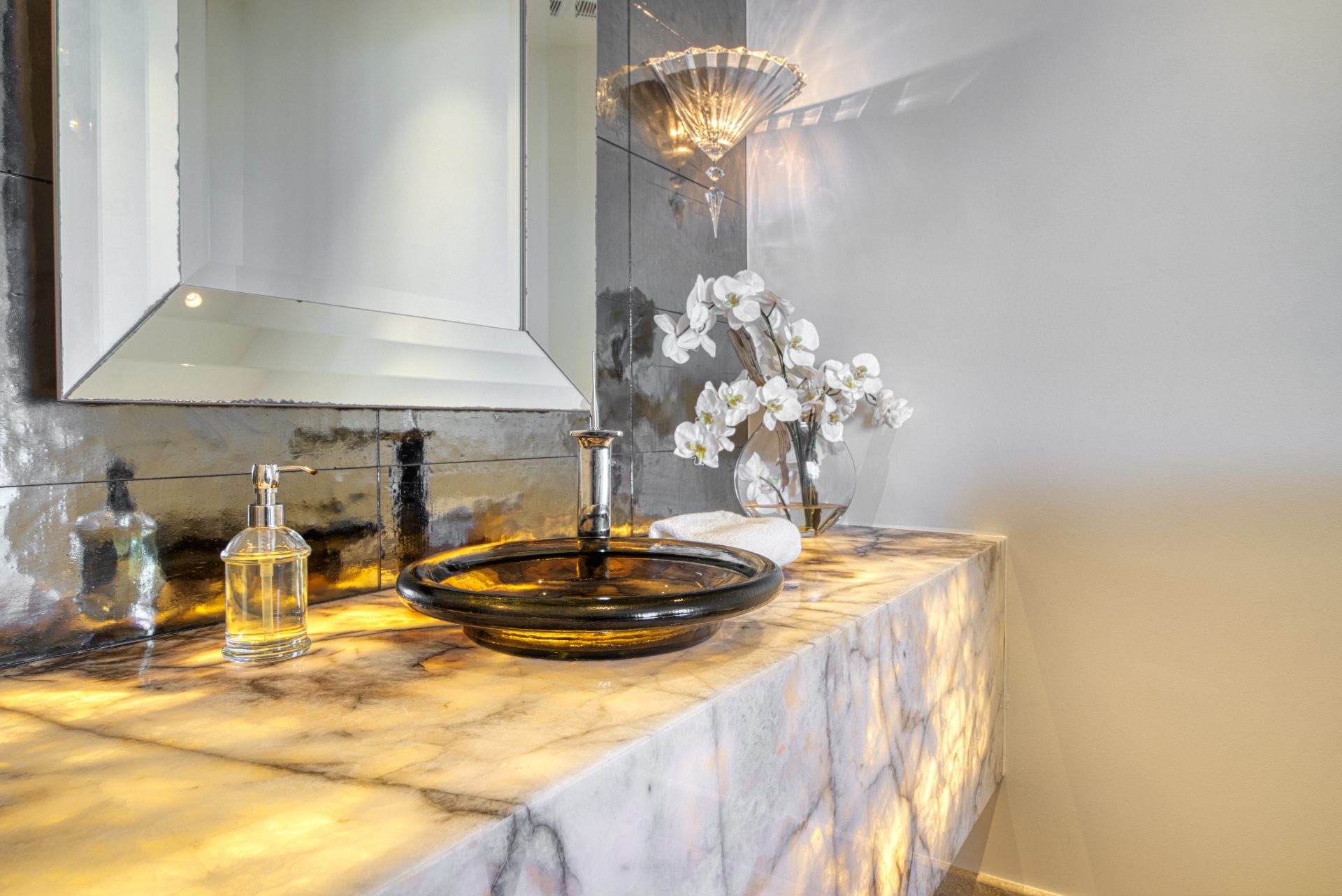 A luxurious bathroom at Rainbow Villa features a marble countertop with a black glass sink and stylish faucet. Next to the sink is a glass soap dispenser, while an elegant arrangement of white orchids adorns the counter. A wall-mounted sconce light illuminates the area, adding to the villa's charm.