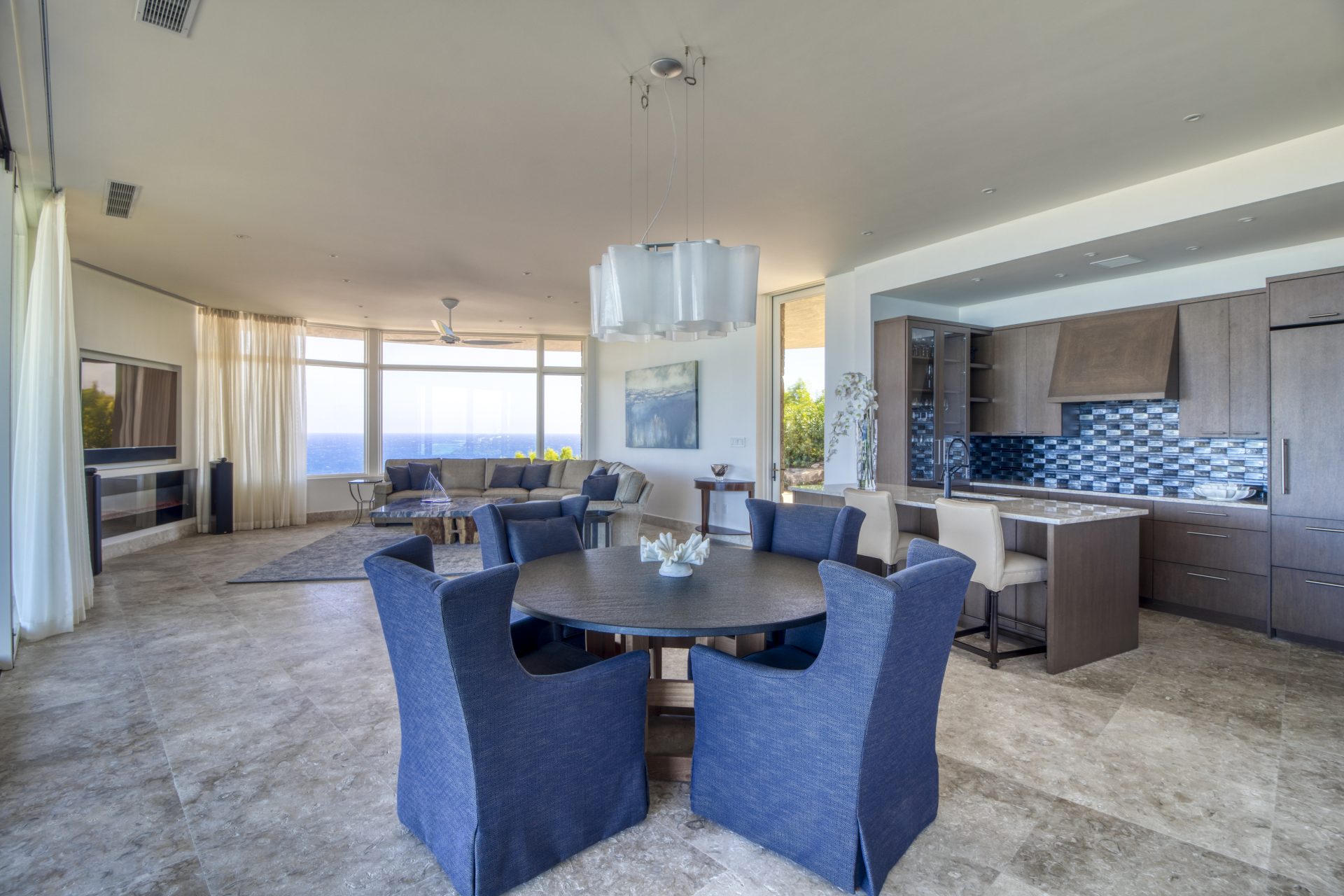 Rainbow Villa boasts a spacious open-concept living area with modern furnishings. The room features a round dining table with blue chairs, a kitchen with dark wood cabinetry and patterned backsplash, and a seating area with a large window providing an ocean view. Marble flooring throughout.