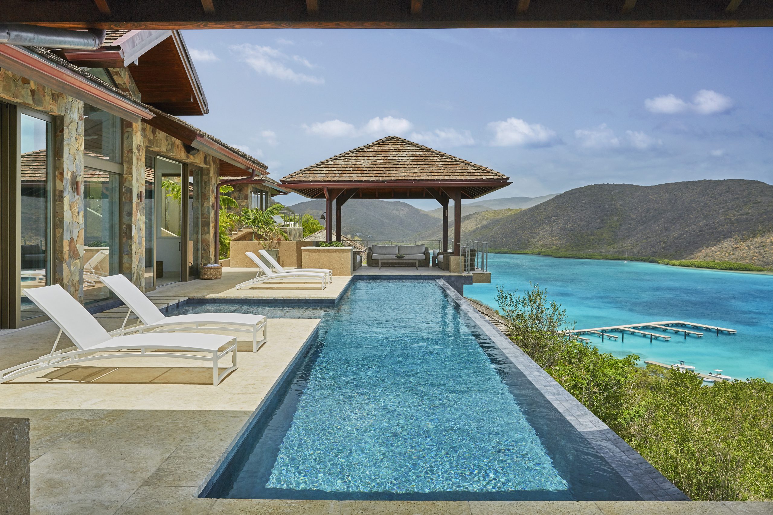 A luxurious infinity pool at an upscale resort overlooks a serene turquoise bay and tree-covered hills. Sun loungers line the pool's edge, and a shaded pavilion sits at the far end. Large windows and a stone facade adorn the adjacent building, giving it the charm of an opulent house by the reef.