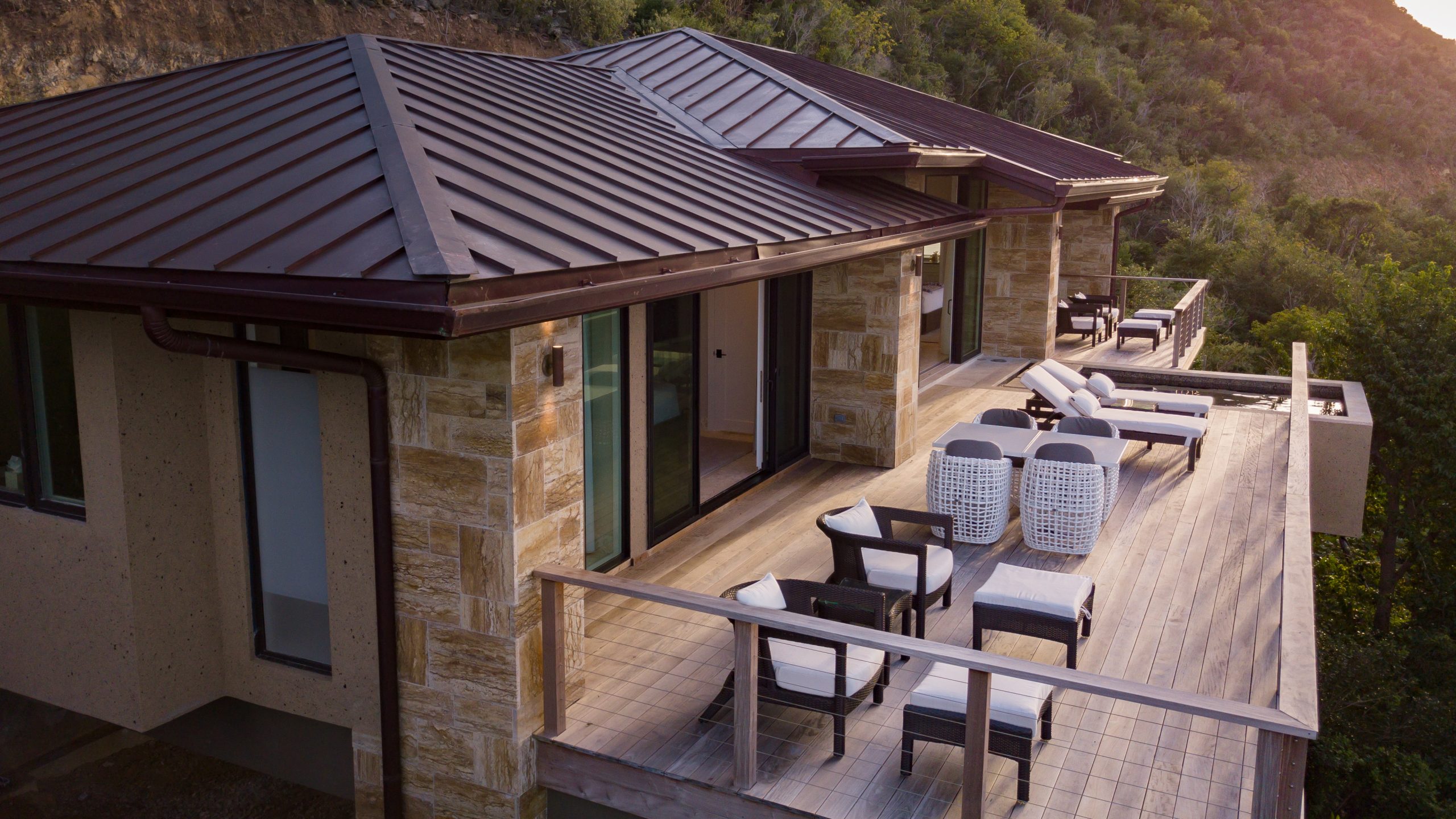 A modern villa with a stone and wood exterior features a large, furnished terrace. The terrace includes several lounge chairs, a sofa, and a small table, all overlooking a lush, green landscape. The house has a metal roof and large windows that capture views of the distant sea.