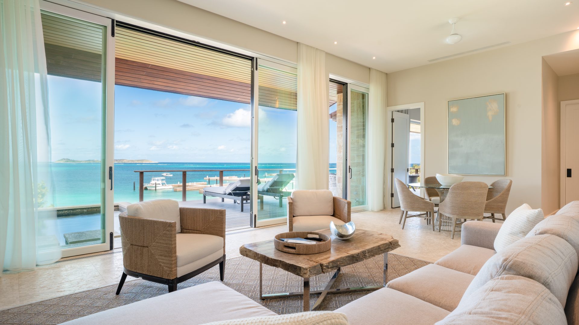 A bright, modern living room opens to a spacious balcony with stunning sea views at Villa Sabella. The room features contemporary furniture, including a white sofa, beige armchairs, and a wooden coffee table. Large sliding glass doors allow natural light to fill the space.