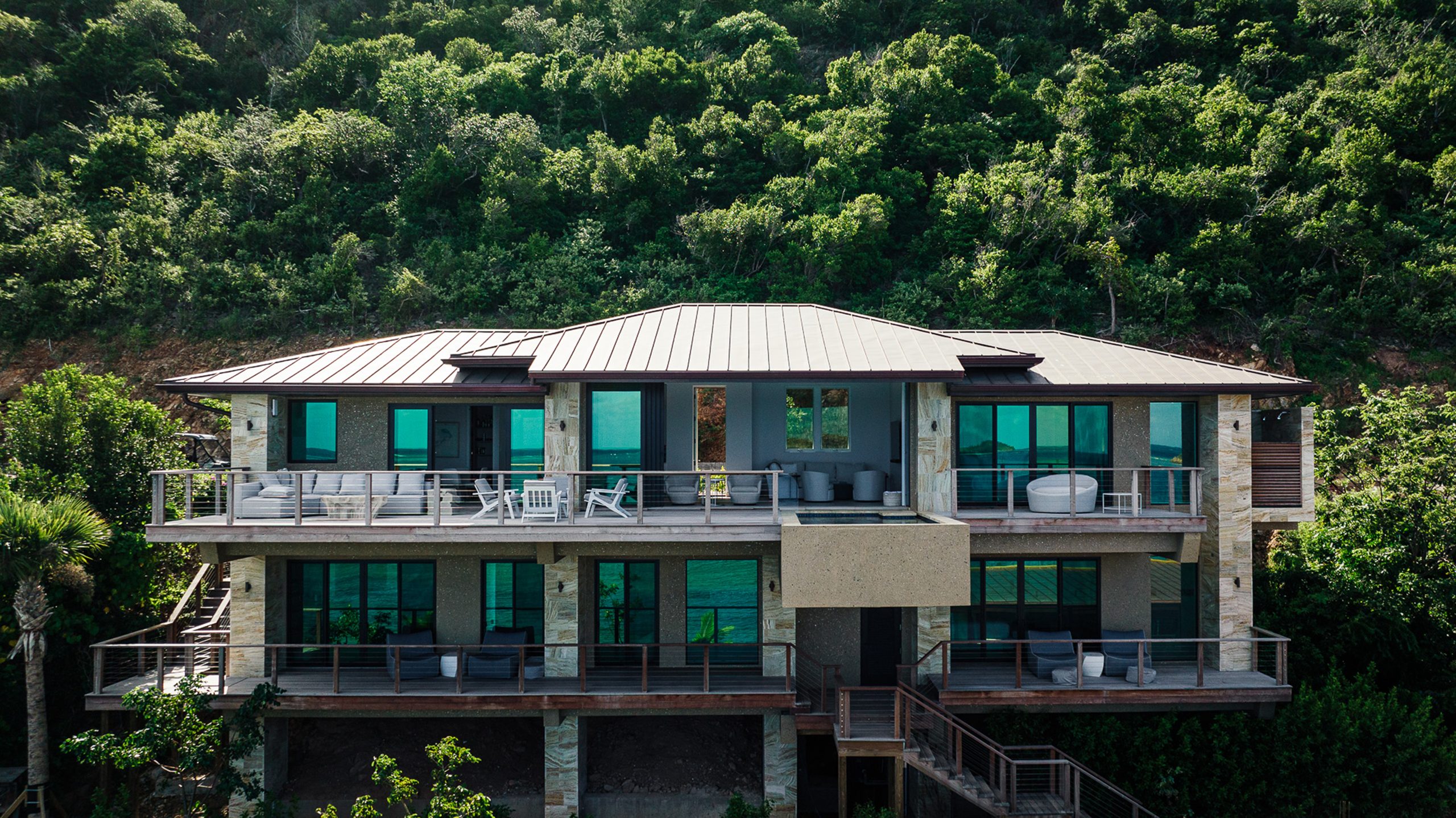 A modern, multi-story villa with large windows and spacious balconies sits nestled against a lush, green hillside. The exterior features clean lines and minimalistic design, surrounded by dense forested vegetation, creating a serene retreat named Shanti.