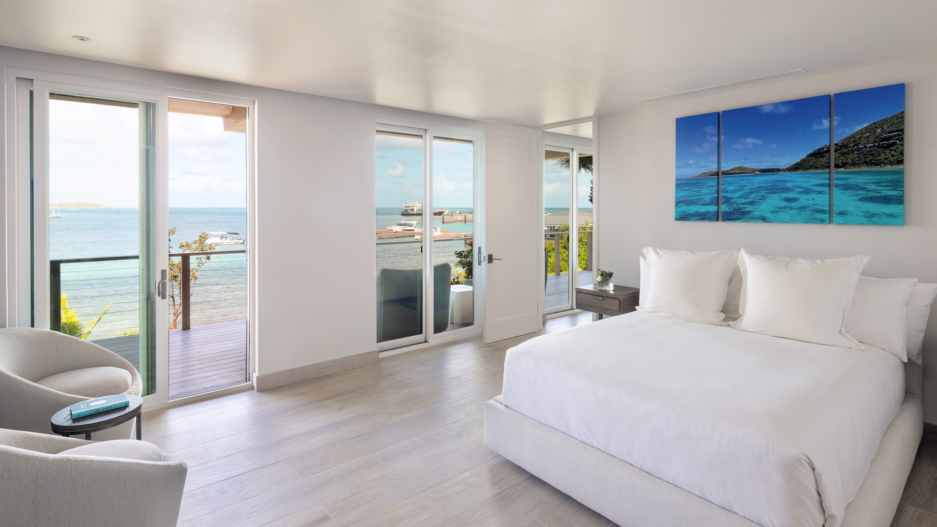 A bright and modern bedroom at Shanti Villa features white walls and furnishings. A large bed is positioned near floor-to-ceiling windows that overlook a balcony with a stunning ocean view. A triptych of ocean-themed artwork hangs above the bed, and there is a cozy chair by the window.