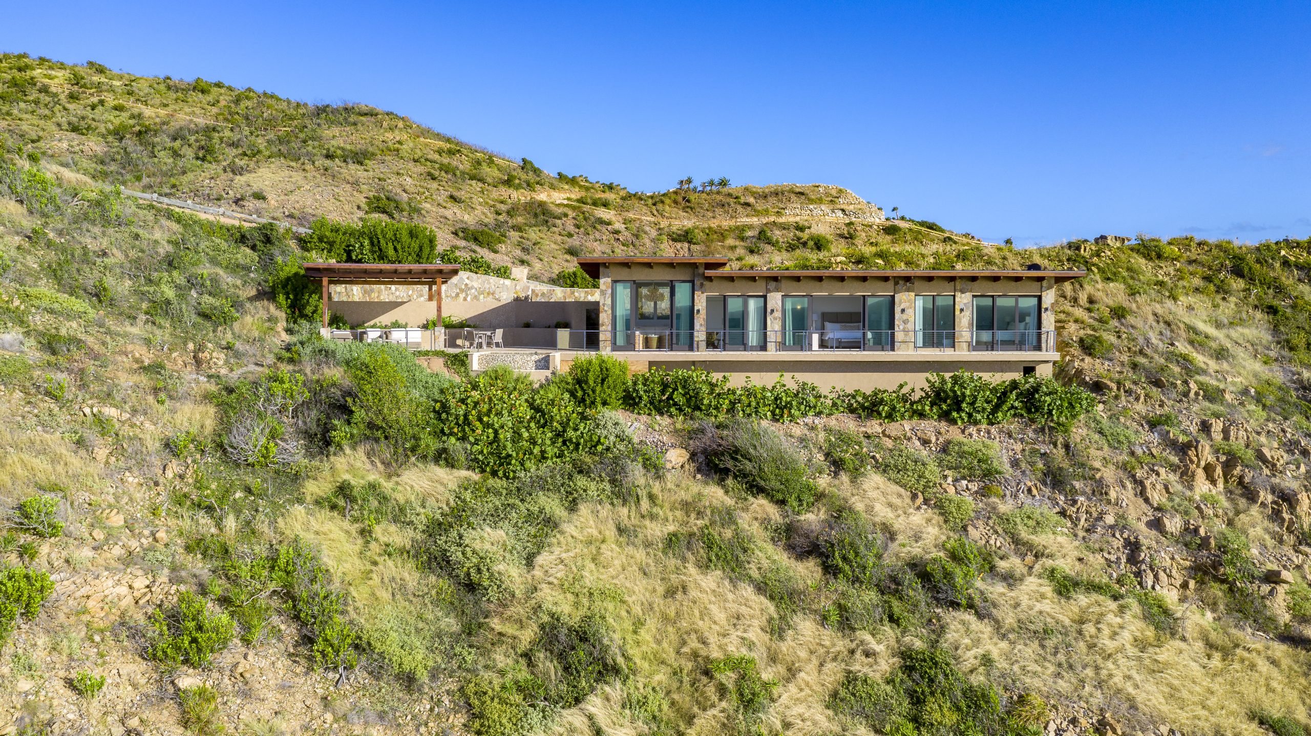 Sol Mate Villa is a contemporary hillside retreat with expansive glass windows and two connected buildings overlooking lush greenery and rugged terrain under a clear blue sky. The property includes outdoor seating areas and offers panoramic views of the surrounding landscape.