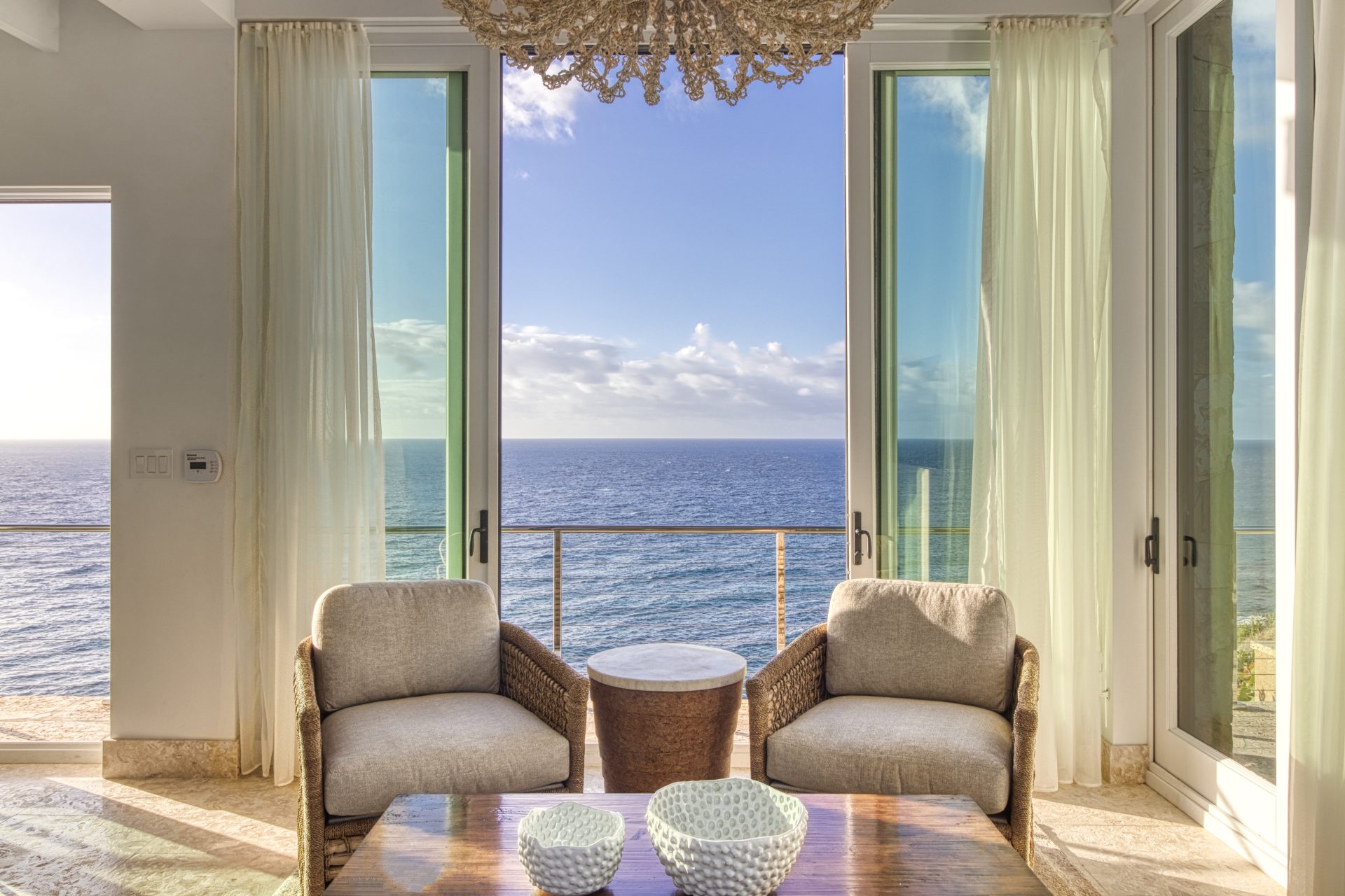 A cozy seating area at Sol Mate Villa features two wicker chairs and a small table facing floor-to-ceiling windows that open up to a stunning ocean view. Sheer curtains frame the windows, letting in natural light, and a decorative chandelier hangs from the ceiling.