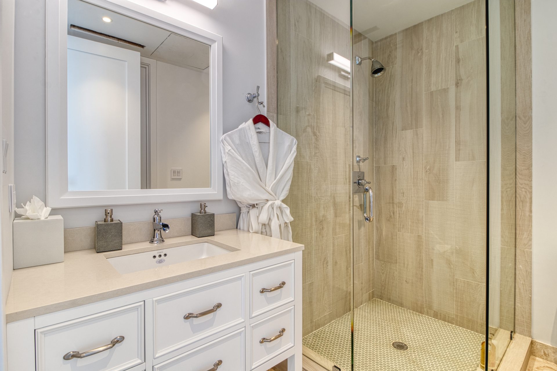 A modern bathroom at Sol Mate Villa featuring a white vanity with a large mirror, sink, and chrome fixtures. A glass-enclosed shower with beige tile walls is to the right, with a white bathrobe hanging on a hook near the door. The design is clean and minimalist.