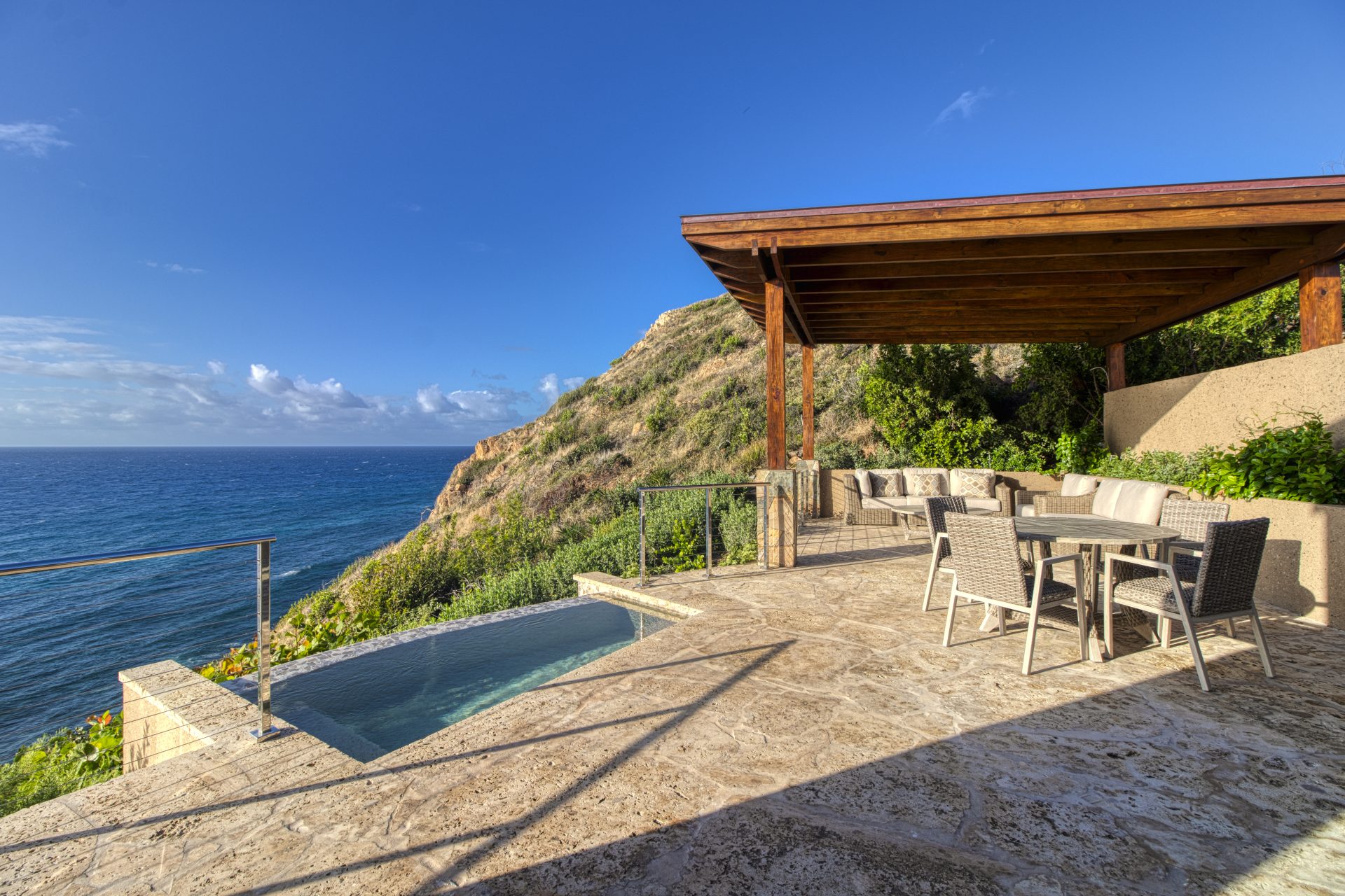 Sol Mate Villa boasts a serene outdoor patio featuring a dining table with four chairs beneath a wooden pergola. Adjacent is a small infinity pool overlooking a scenic coastline. The backdrop includes clear blue skies, a rocky hillside, and the expansive ocean.